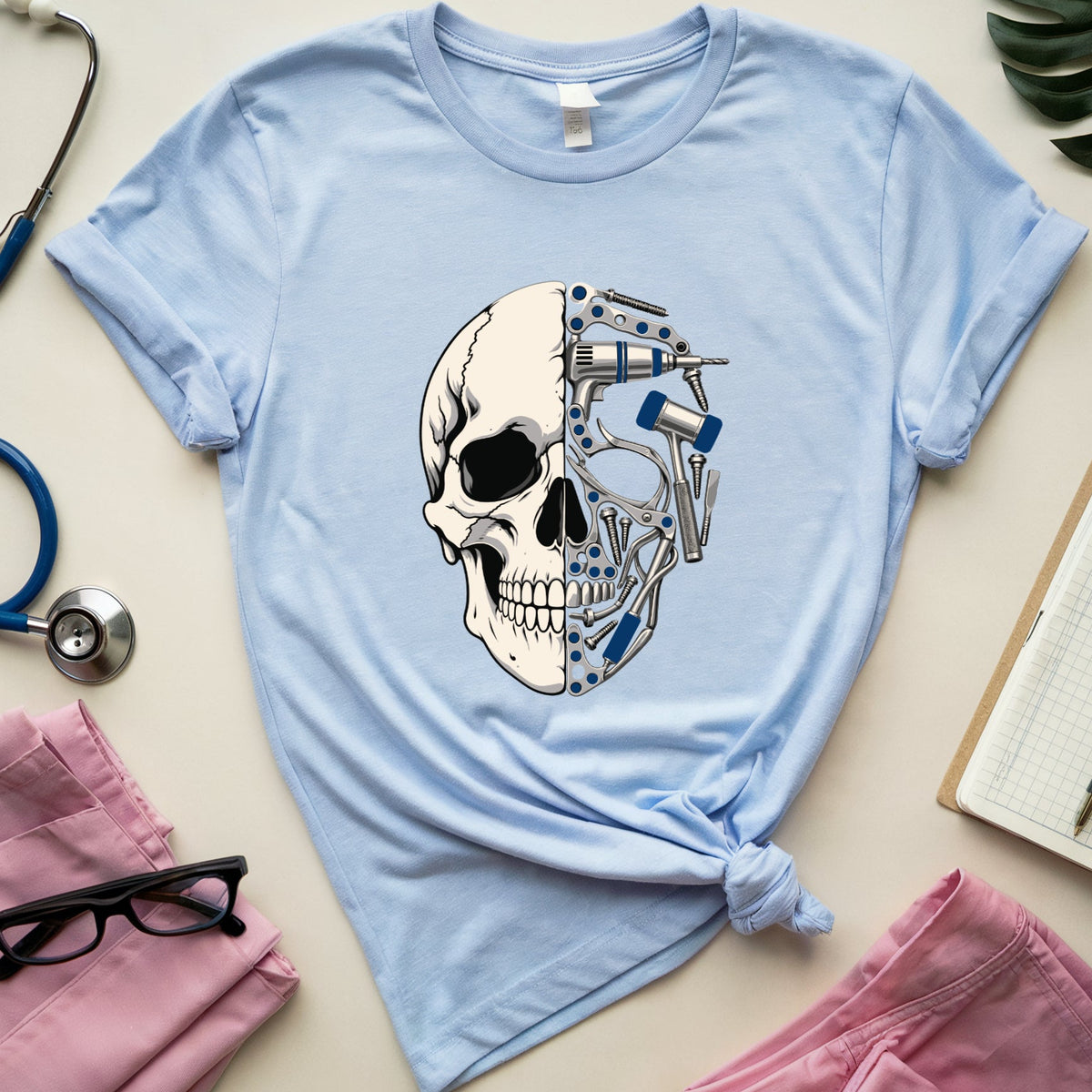 A light blue graphic t-shirt featuring a split skull design with medical tools on one side and a bone structure on the other. The t-shirt is styled in a flat lay with medical-themed props including a blue stethoscope, pink scrubs, glasses, a notebook, and a green leaf on a beige background.