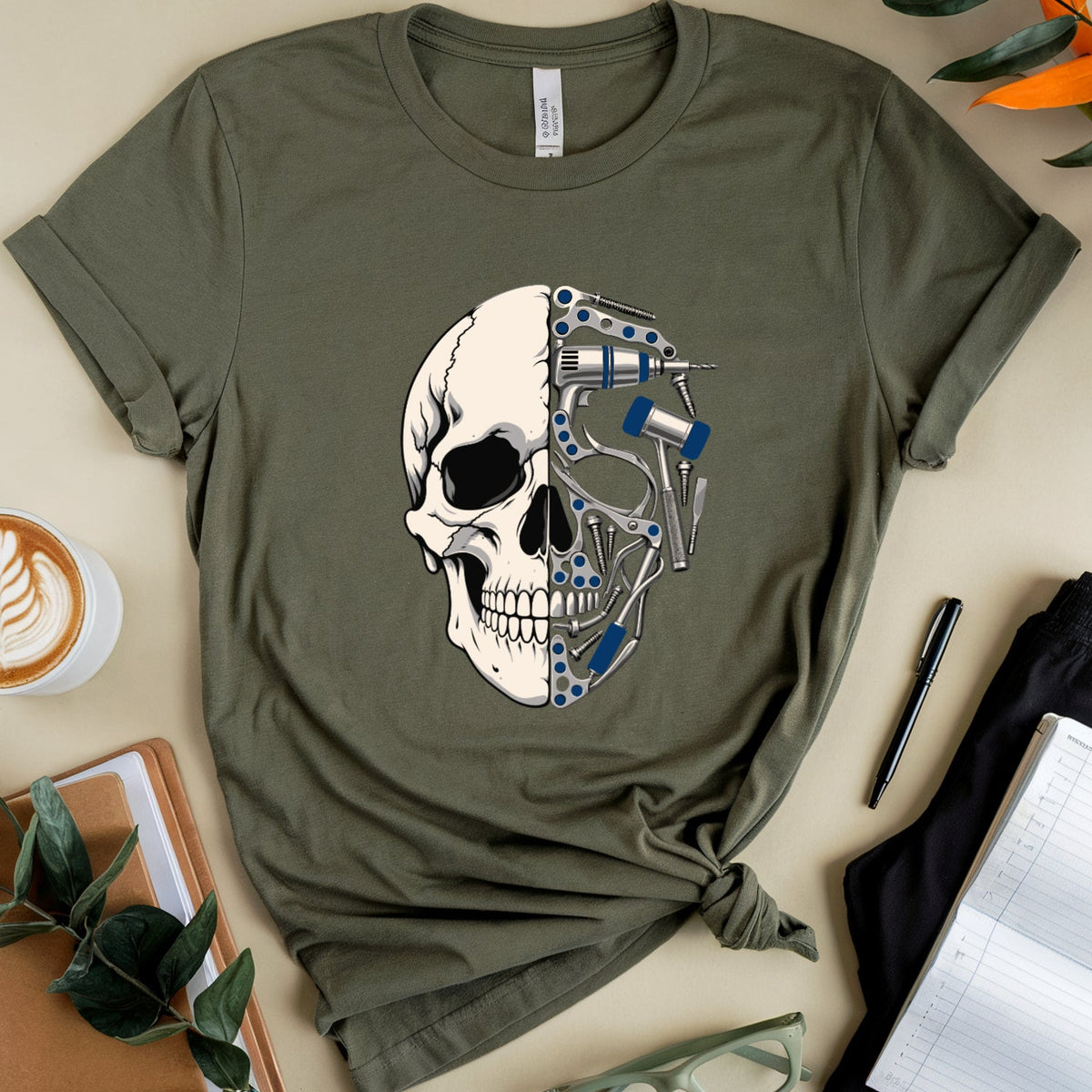 An olive green short-sleeve t-shirt with a graphic of a human skull merged with robotic mechanical parts, styled in a flat lay with a coffee cup, notebook, glasses, and leaves on a beige background.