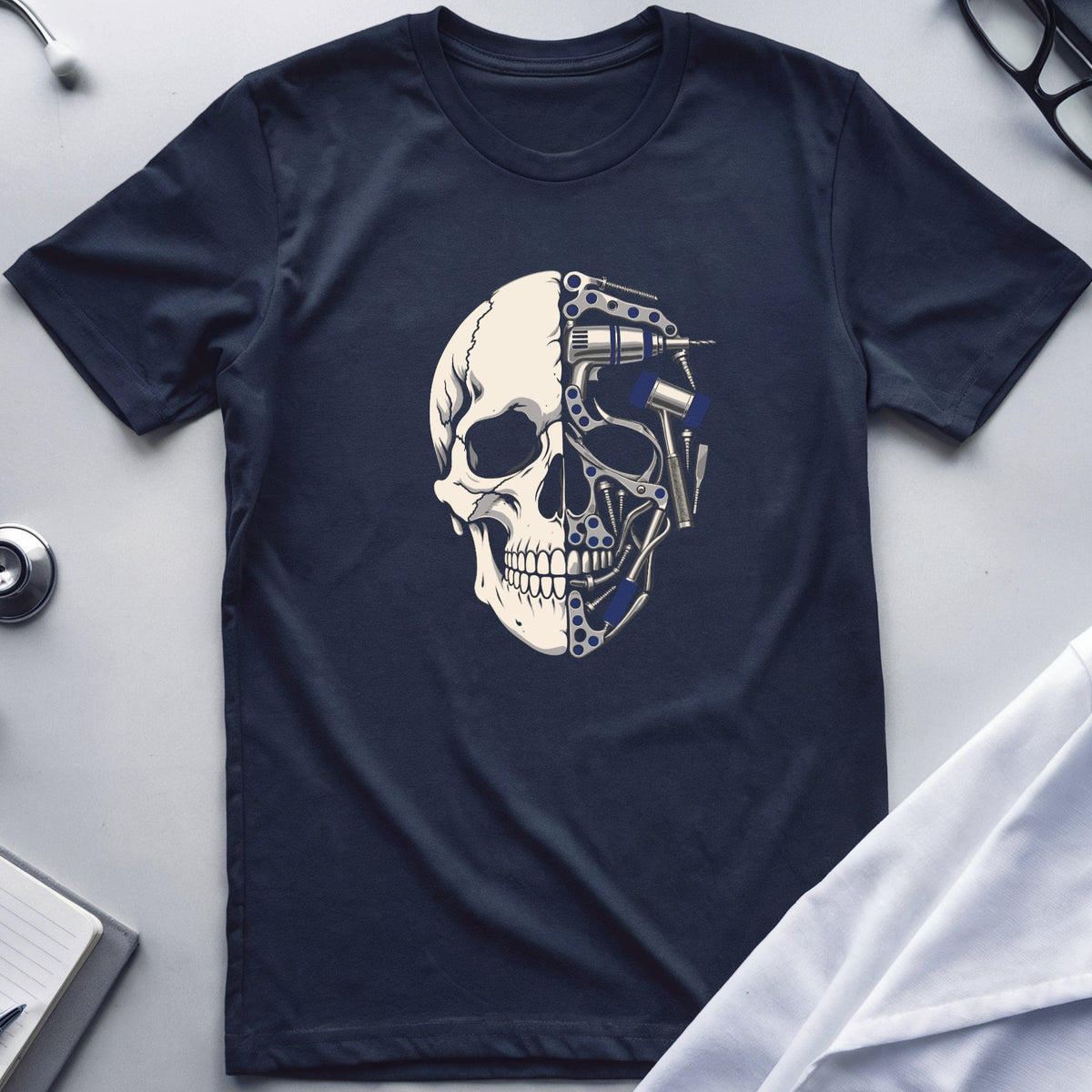 A navy blue short-sleeve t-shirt with a graphic of a split human skull, one half showing a detailed bone structure and the other half showing mechanical gears and circuitry. The shirt is presented in a flat-lay arrangement on a light gray surface, surrounded by medical-themed props including a stethoscope, glasses, a notebook, and a white lab coat.