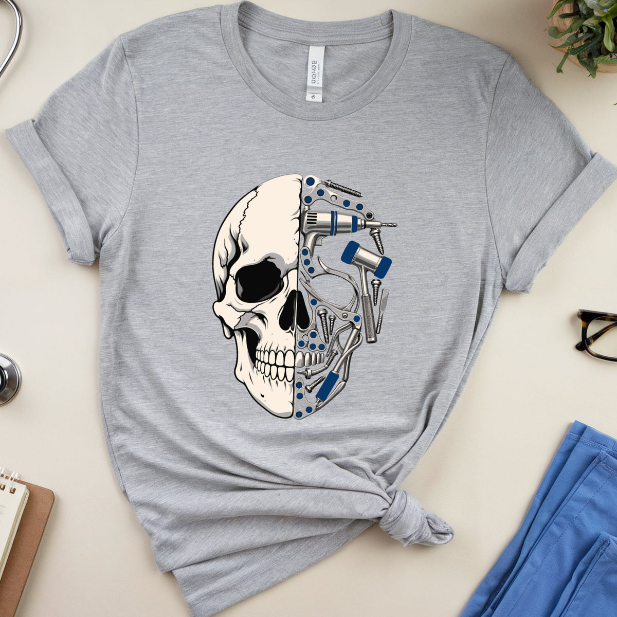A heather gray short-sleeve t-shirt with a graphic of a human skull where one half is replaced by medical tools, styled in a flat lay with a stethoscope, notebook, glasses, and blue scrubs on a beige background.