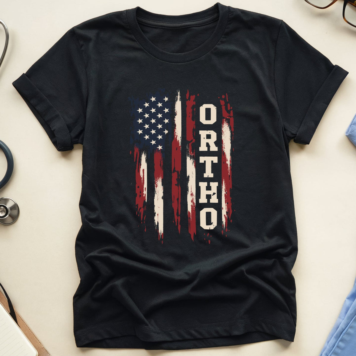 A black short-sleeve T-shirt laid flat on a light beige surface, featuring a distressed American flag graphic with the word 'ORTHO' printed vertically in white over the stripes. Surrounding props include a stethoscope, eyeglasses, a notebook with pen, and light blue scrubs.