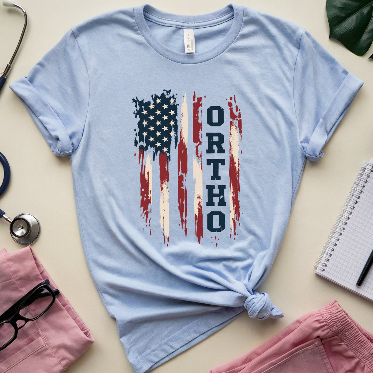 A light blue short-sleeve T-shirt with a distressed American flag graphic and the word 'ORTHODOX' printed vertically, styled in a flat lay with pink scrubs, a stethoscope, glasses, a notebook, and a plant leaf on a light beige background.