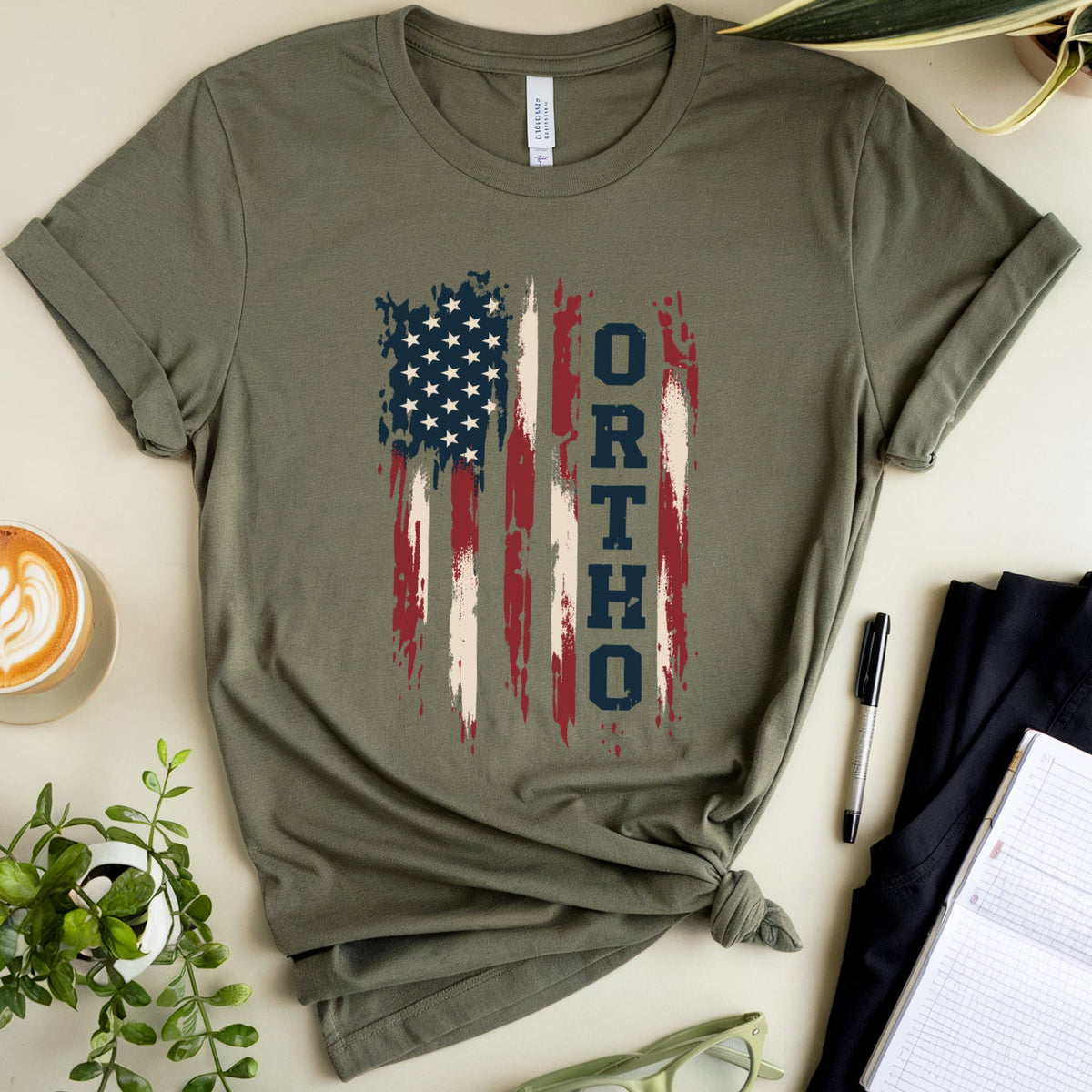 An olive green short-sleeve t-shirt with a distressed American flag graphic and the word 'ORTHO' printed vertically, styled in a flat lay with a coffee cup, plants, glasses, a pen, and a notebook on a light beige background.