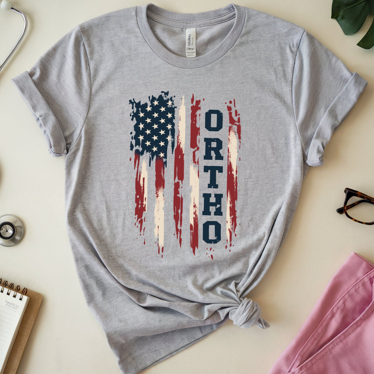 A heather gray short-sleeve T-shirt laid flat on a light beige surface, styled with a stethoscope, notebook, eyeglasses, green leaves, and pink fabric around it. The shirt has a distressed American flag graphic with the word 'ORTHO' printed vertically in blue.