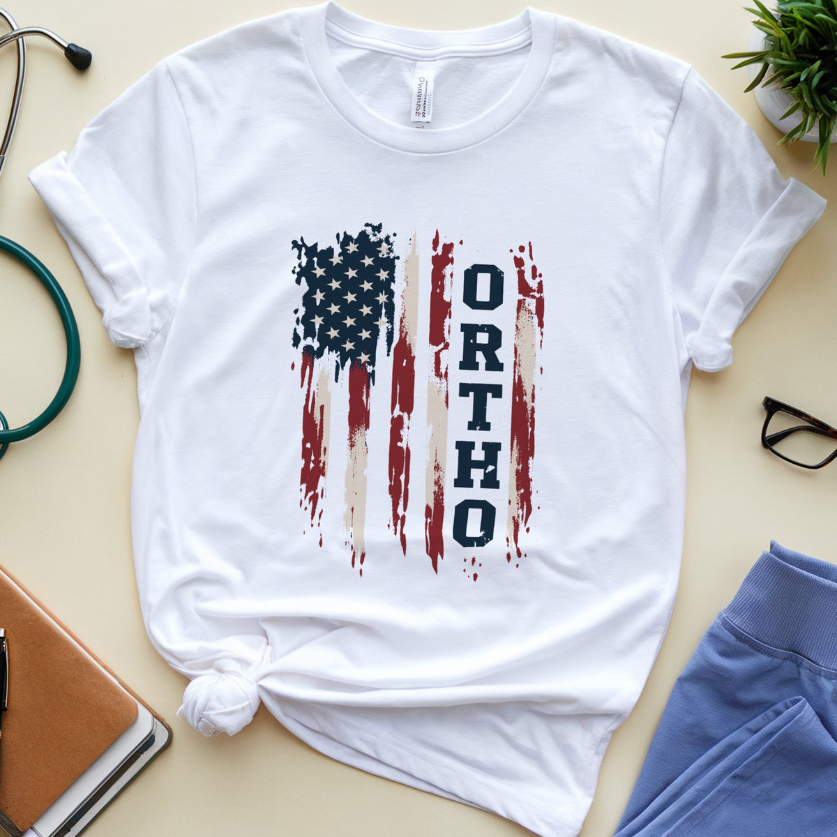 A white short-sleeve T-shirt laid flat on a light beige surface, styled with a knotted hem and a distressed American flag graphic featuring the word 'ORTHODOX' in vertical navy text. Surrounding props include a stethoscope, notebook, pen, glasses, blue pants, and a small potted plant.