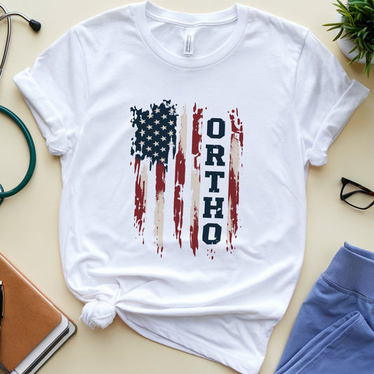 A white short-sleeve T-shirt laid flat on a light beige surface, styled with a knotted hem and a distressed American flag graphic featuring the word 'ORTHODOX' in vertical navy text. Surrounding props include a stethoscope, notebook, pen, glasses, blue pants, and a small potted plant.