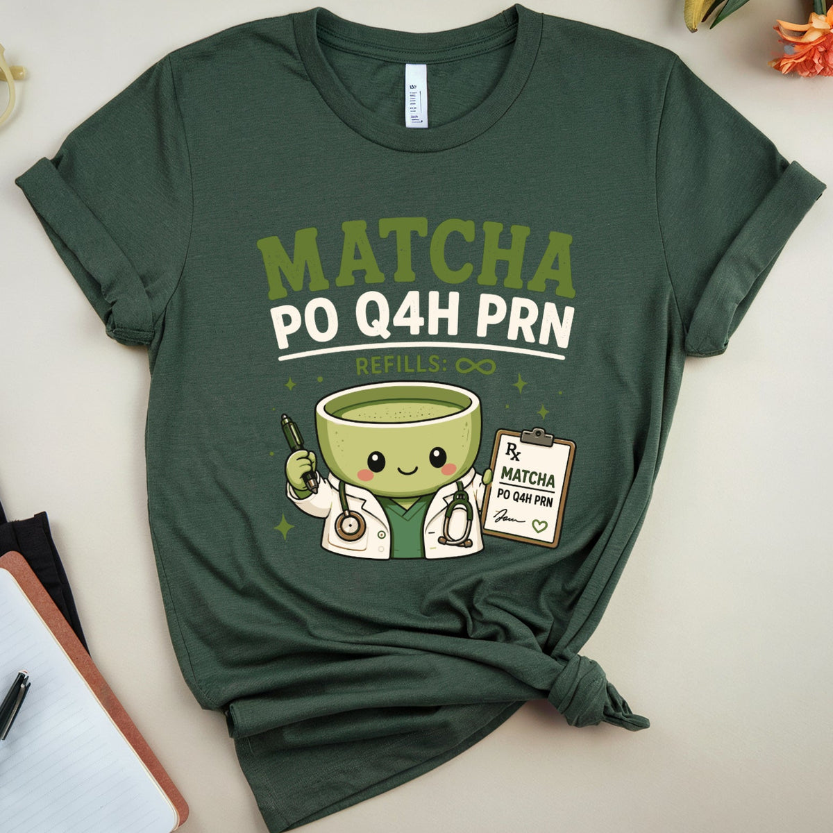 A dark green graphic t-shirt featuring a cute matcha green tea character and the text 'MATCHA PO Q4H PRN' in a styled flat-lay arrangement with a notebook, glasses, and flowers.