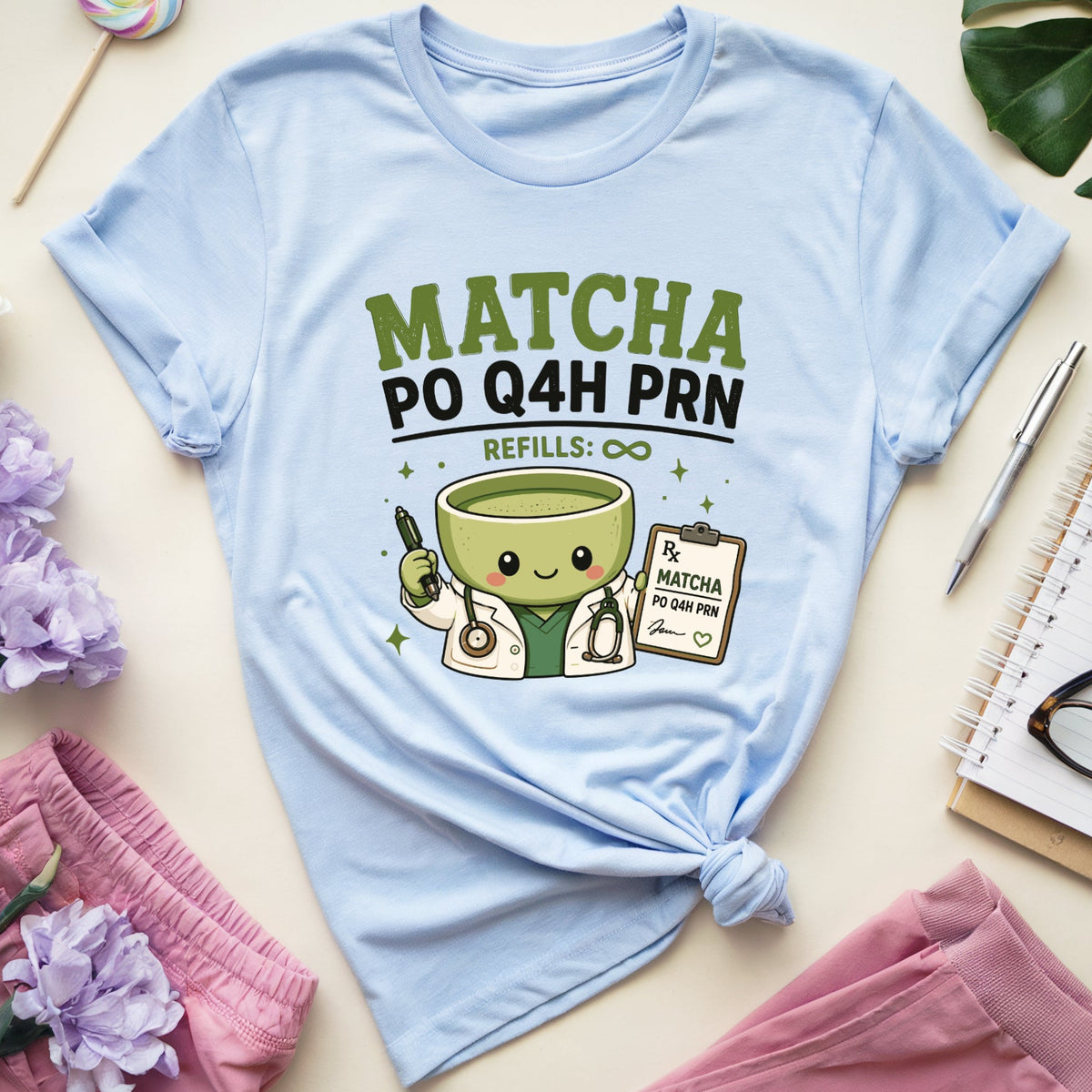 A light blue graphic t-shirt featuring a cute green matcha cup character and the text 'MATCHA PO Q4H PRN REFILLS: ©' in green and black. The t-shirt is styled in a flat lay with pink shorts, purple flowers, a lollipop, a green leaf, a pen, and an open notebook on a light beige background.
