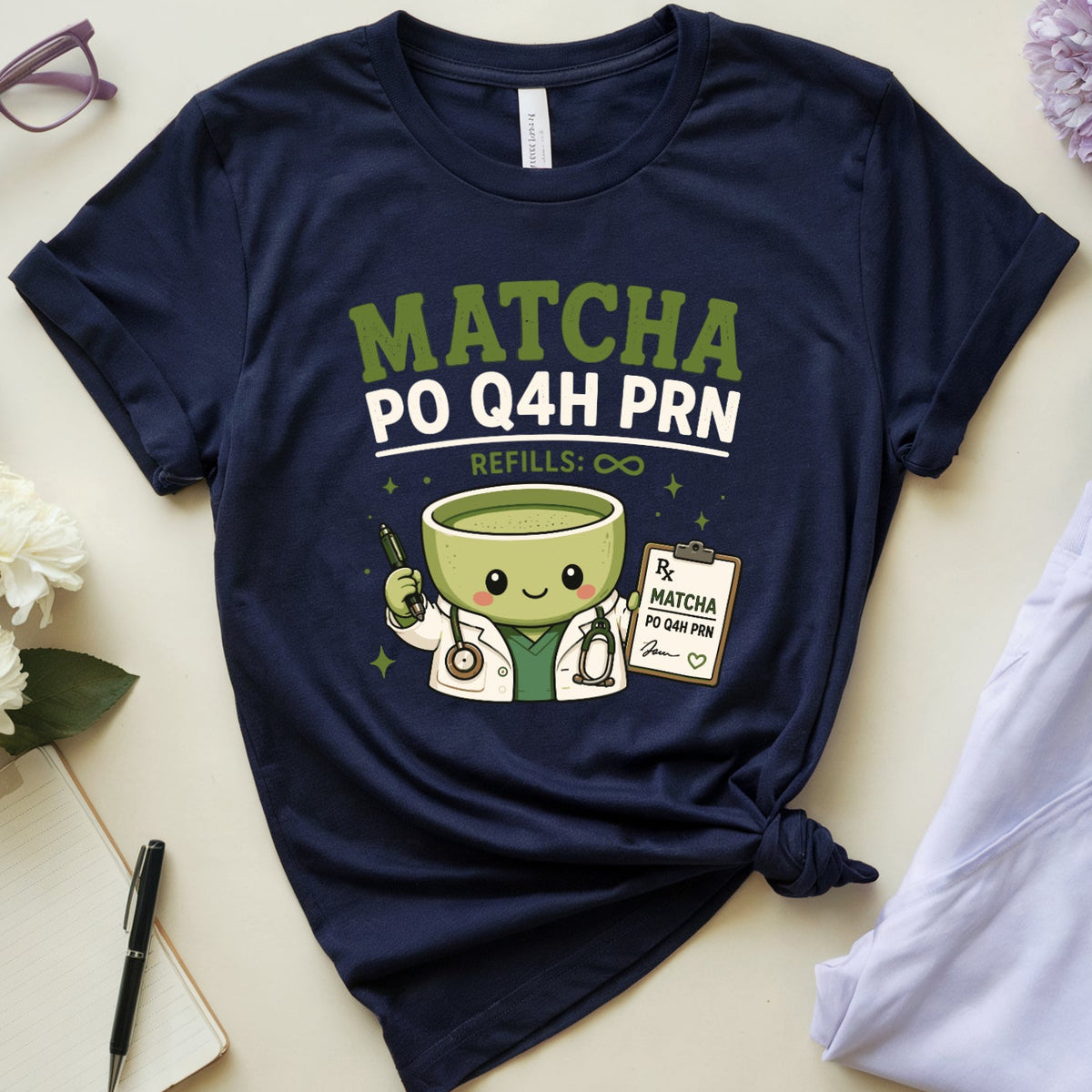 A navy blue graphic t-shirt styled in a flat lay with a knotted hem, featuring a cute matcha tea cup doctor design with the text 'MATCHA PO Q4H PRN'. The shirt is surrounded by a mug of coffee, folded light purple pajama pants, a notebook, glasses, and flowers on a light beige background.
