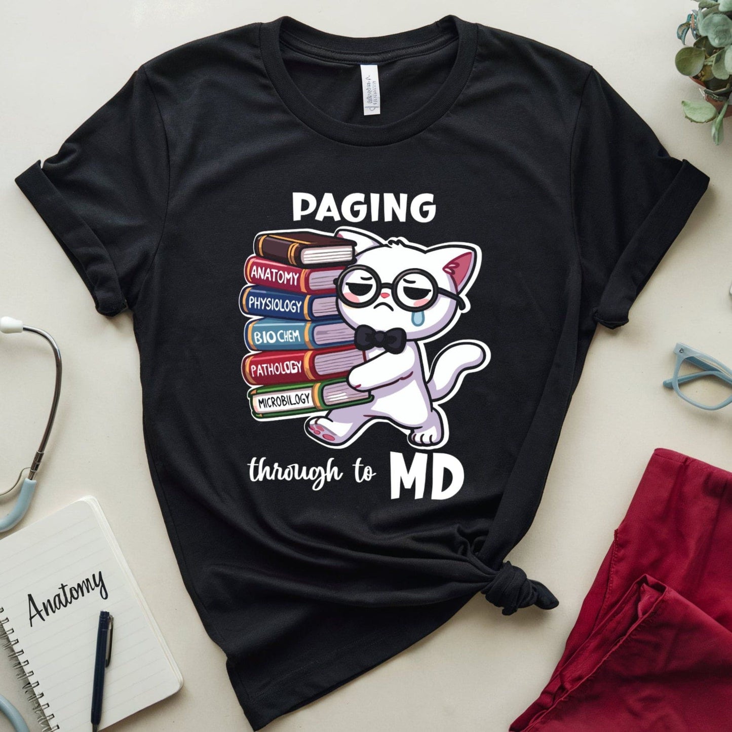 Black t-shirt with a cat graphic and text 'Paging through to MD' on a light surface with medical-themed items.