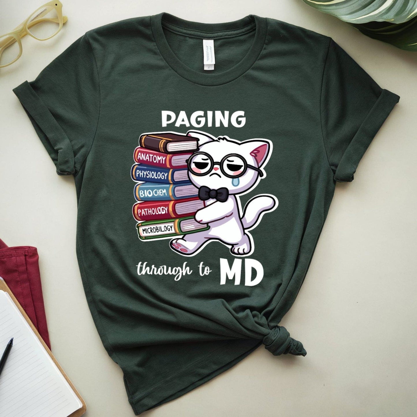 Green t-shirt with a cat graphic and text 'Paging through to MD' on a light surface with books and glasses.