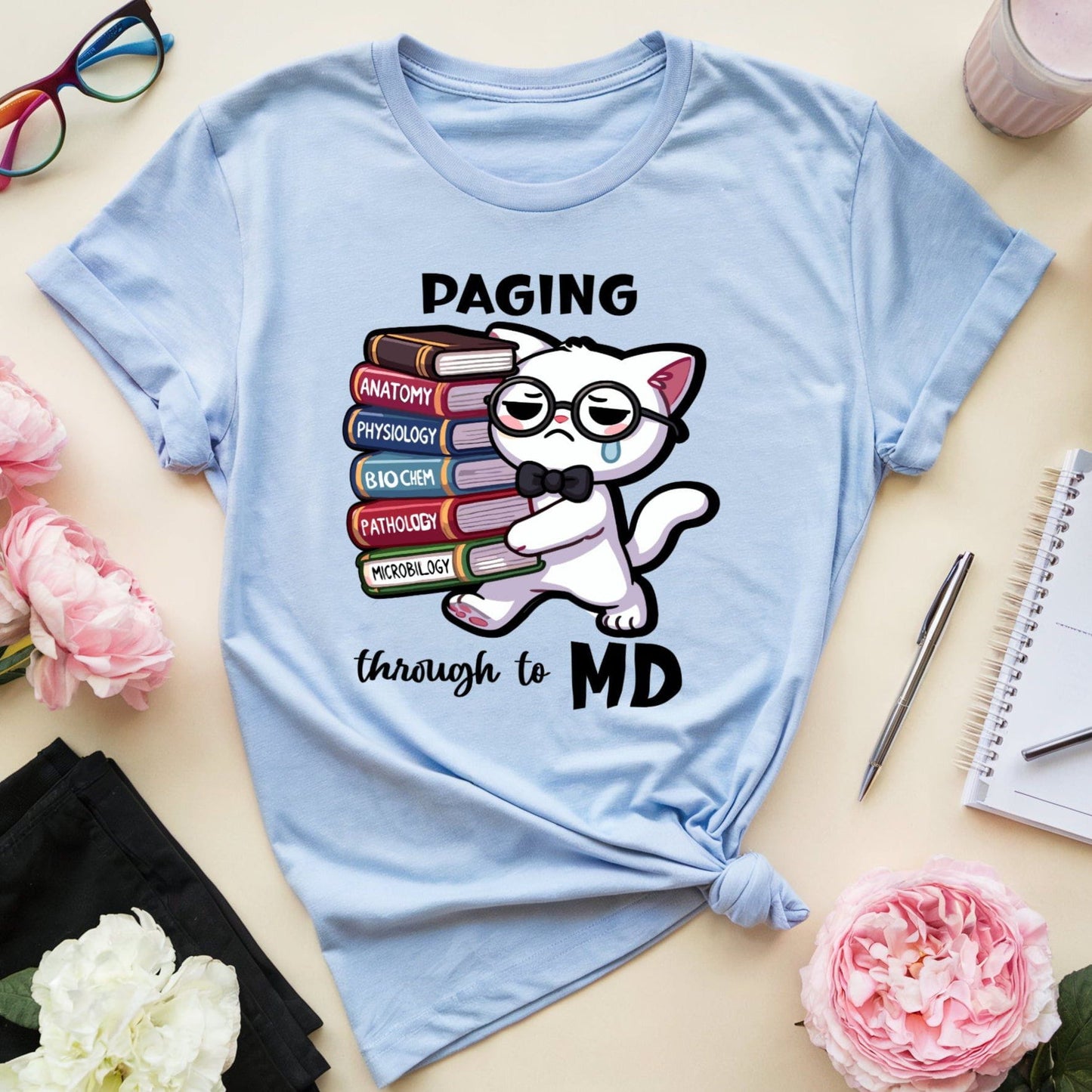 Light blue t-shirt with a cat graphic and text on a table with flowers and glasses.