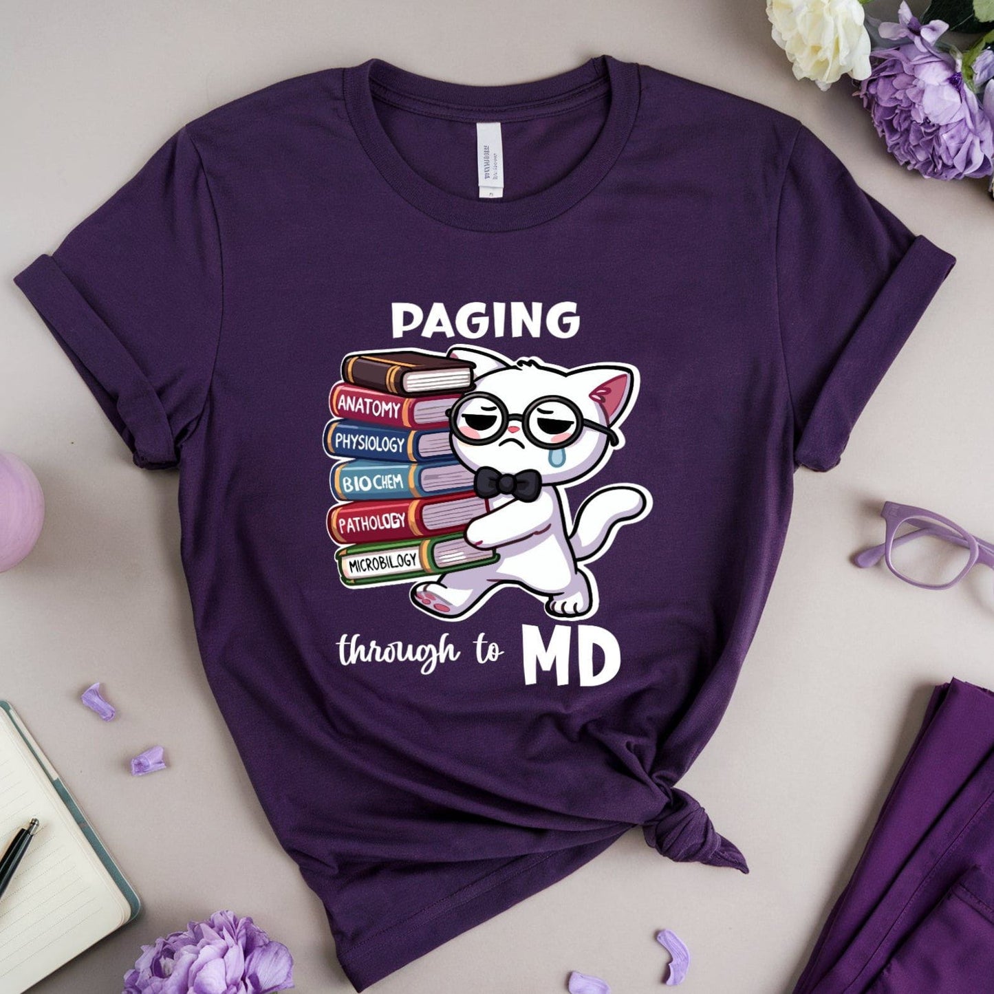 Purple t-shirt with a cat graphic and text 'Paging through to MD' on a light gray surface with flowers and glasses.