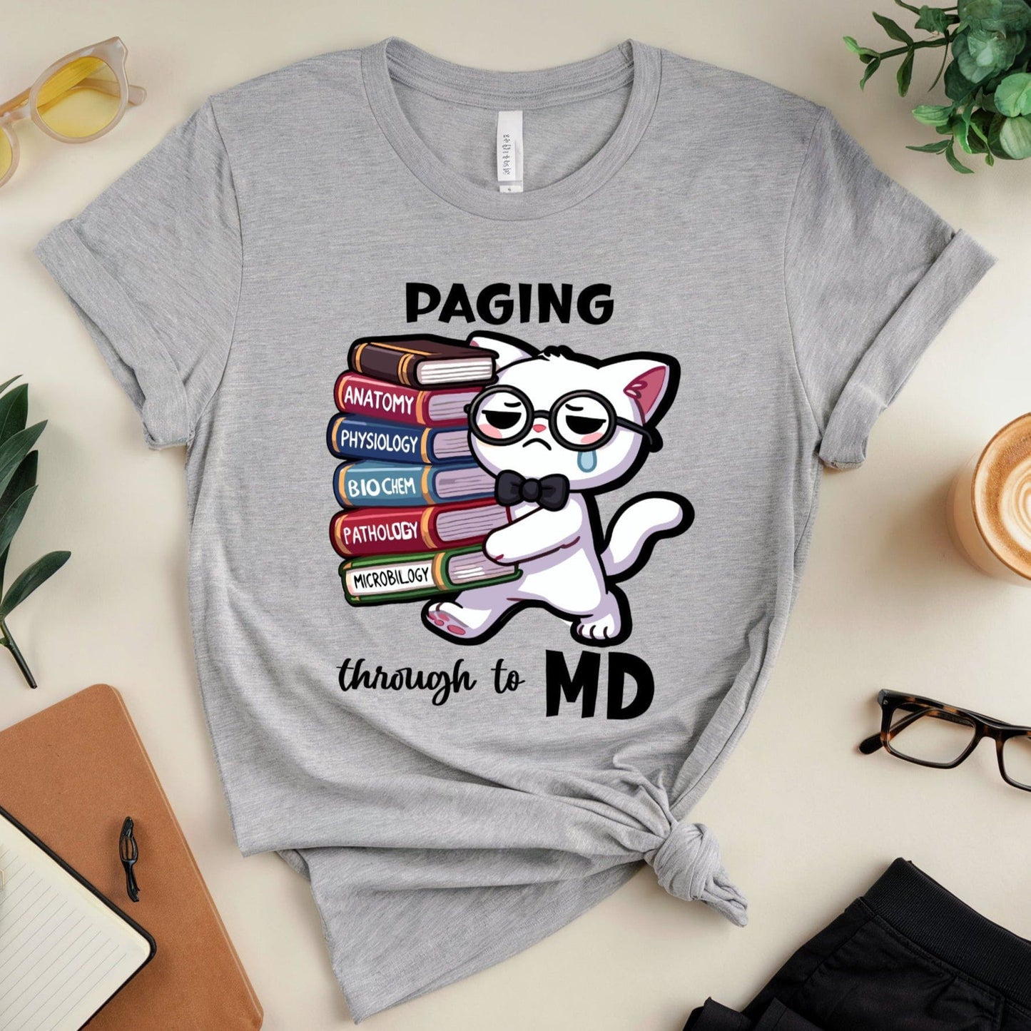 Gray t-shirt with a cat graphic and text 'Paging through to MD' on a desk with accessories.