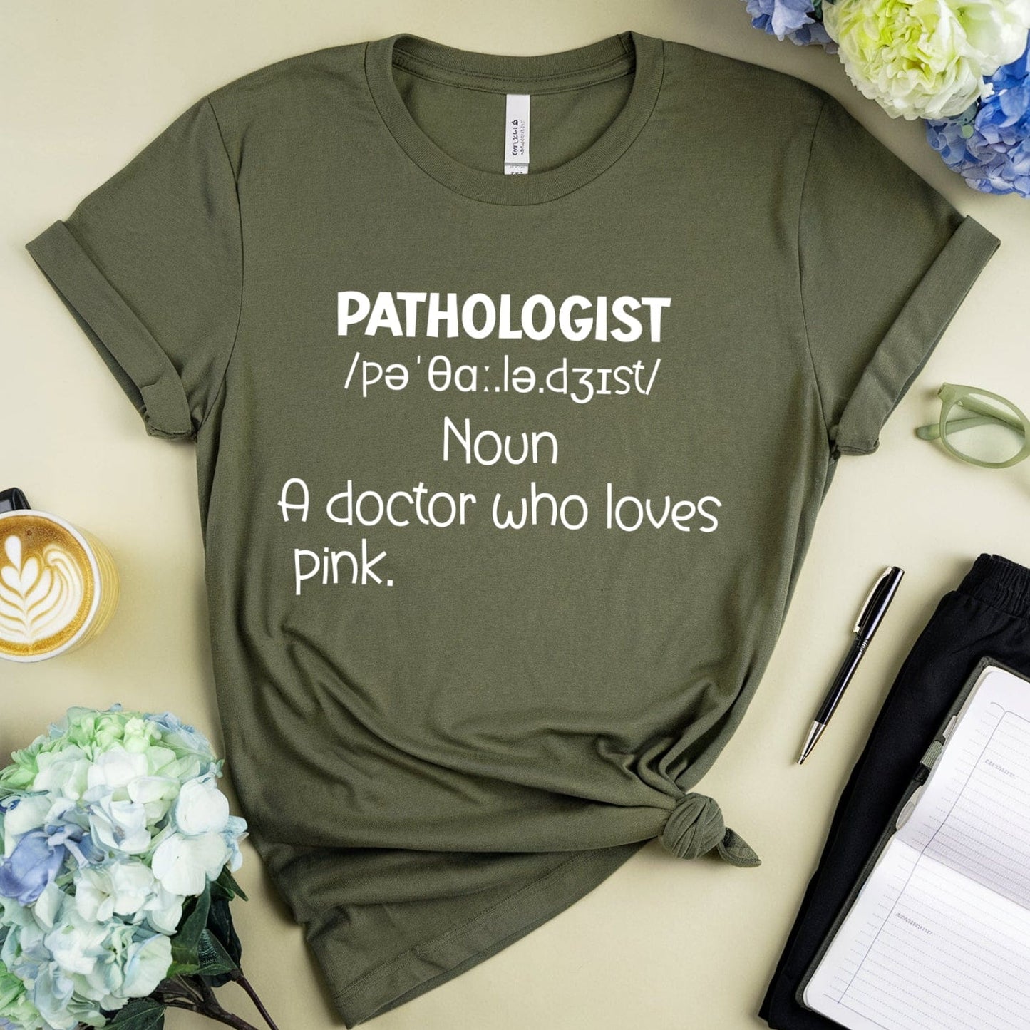 Green t-shirt with 'PATHOLOGIST' definition text, surrounded by flowers and stationery on a beige surface.
