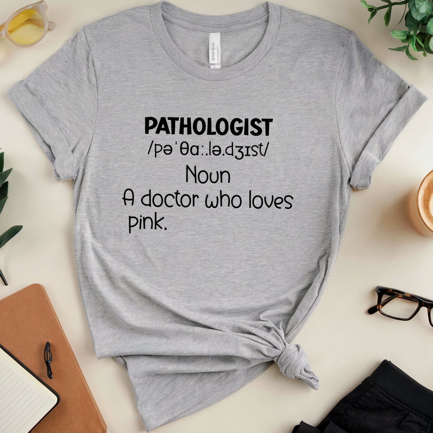 Gray t-shirt with 'PATHOLOGIST' definition text on a desk with office supplies.