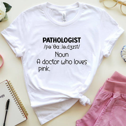 White t-shirt with 'PATHOLOGIST' definition text on a light surface with pink pants and flowers.