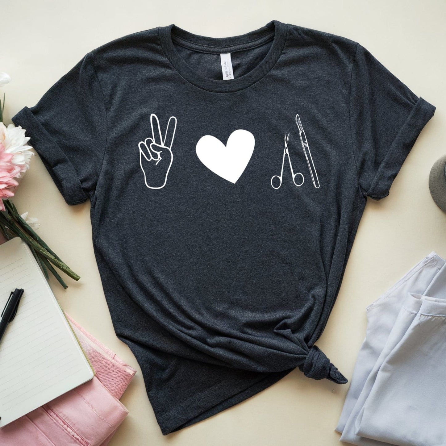 Black t-shirt for surgeons with peace sign, heart, and scissors design on a light surface with jeans, coffee, and flowers.