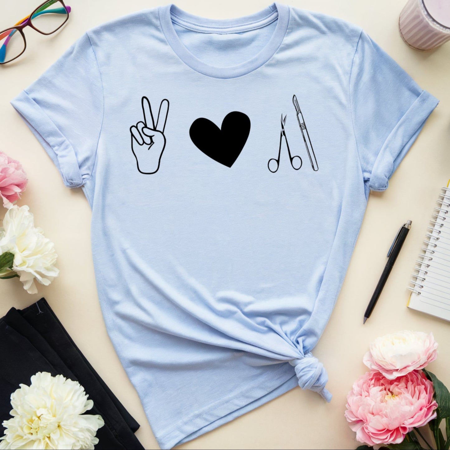 Light blue t-shirt for surgeons with peace sign, heart, and scissors design on a table with flowers and glasses.