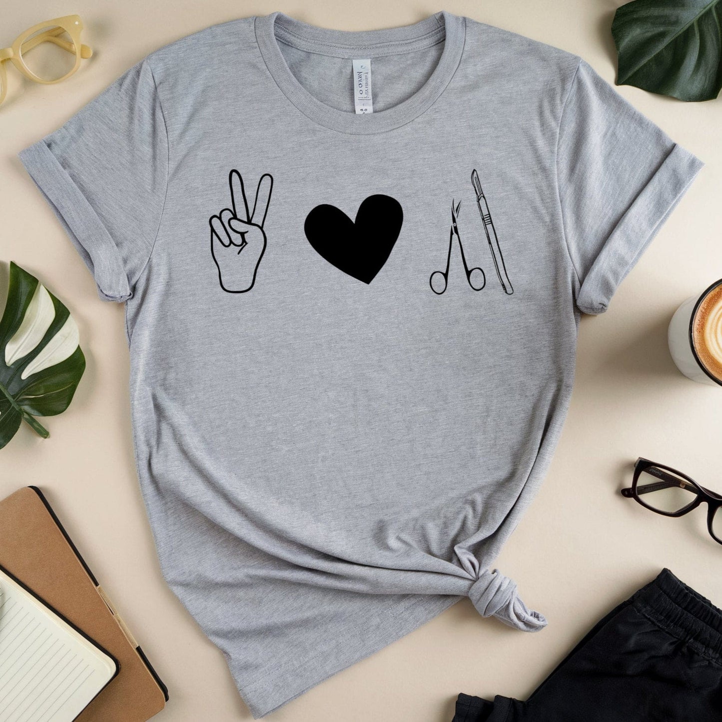 Gray t-shirt for surgeons with peace sign, heart, and scissors design on a beige surface with accessories.