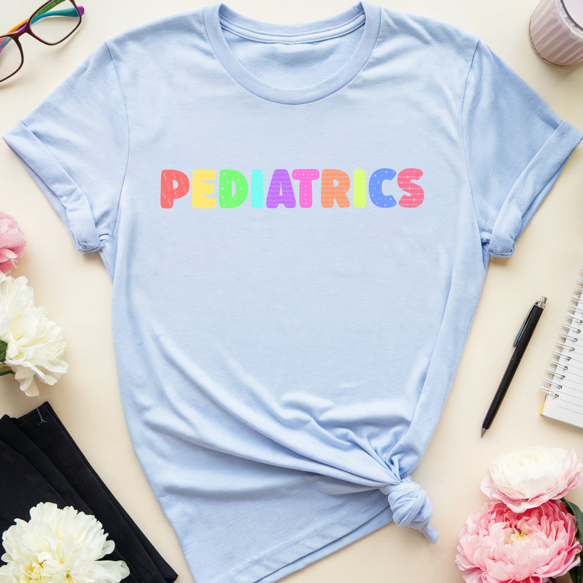 A light blue short-sleeve T-shirt with the word 'PEDIATRICS' printed across the chest in rainbow-colored letters, styled in a flat lay with flowers, glasses, a notebook, and other desk accessories on a light beige background.