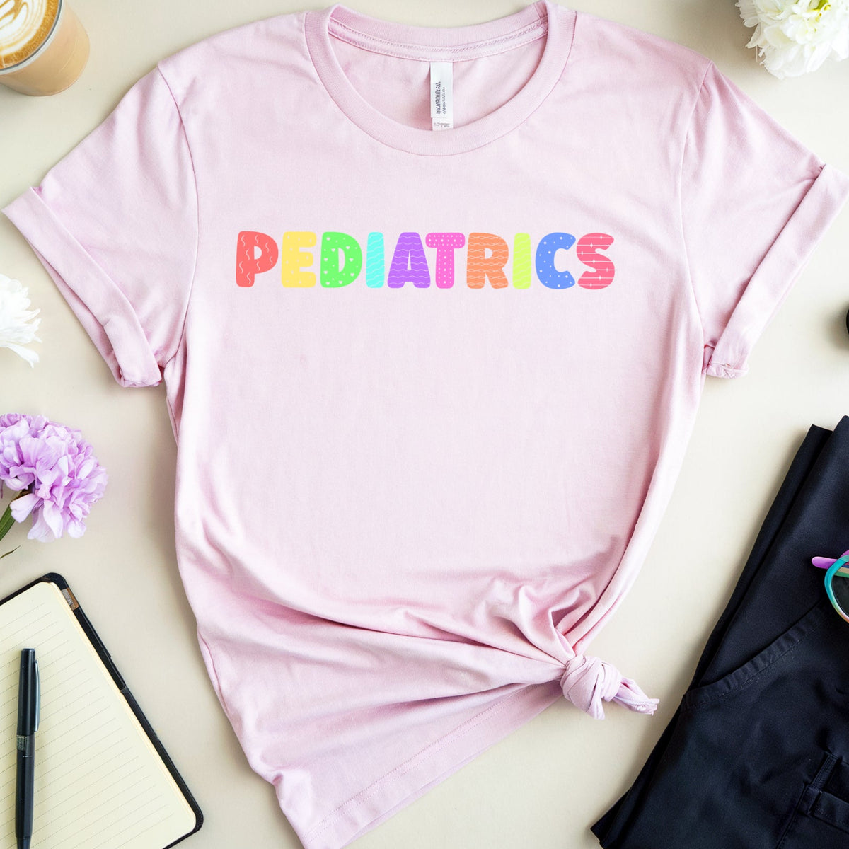 A light pink crewneck t-shirt with the word 'PEDIATRICS' printed across the chest in colorful rainbow letters. The t-shirt is styled in a flat lay with a knot at the bottom, surrounded by a coffee cup, flowers, a notebook, a pen, folded navy pants, and rainbow-framed glasses on a light beige background.