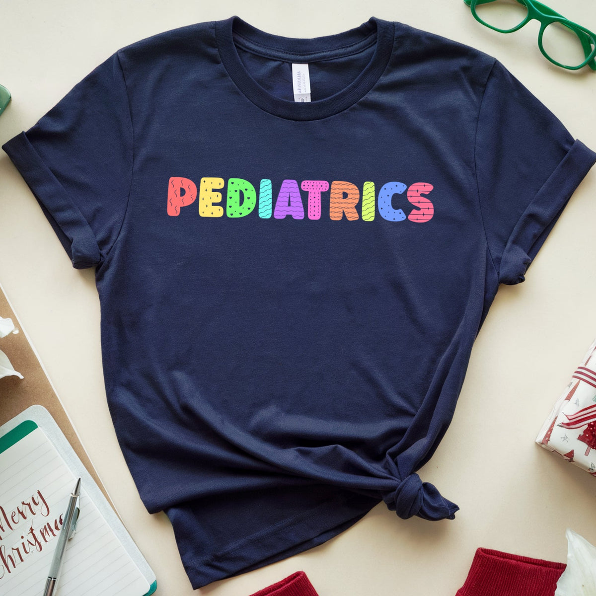 A navy blue t-shirt with the word 'PEDIATRICS' printed across the chest in colorful, rainbow-like letters. The shirt is styled in a flat-lay arrangement with Christmas-themed props including a gift box, a notebook, red pants, and green glasses on a light beige background.