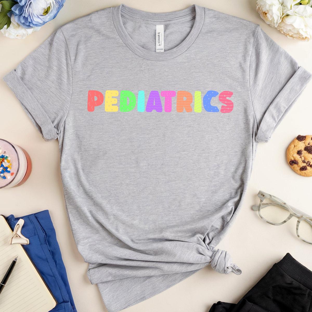 A heather gray short-sleeve T-shirt with the word 'PEDIATRICS' printed across the chest in colorful bubble letters, styled in a flat lay with flowers, a drink, glasses, a cookie, and clothing accessories on a light beige background.