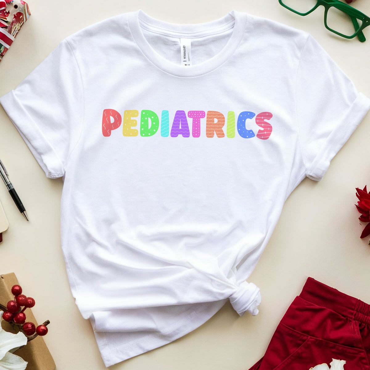 A white short-sleeve T-shirt with the word 'PEDIATRICS' printed across the chest in rainbow-colored letters, styled in a flat lay with holiday gifts, red shorts, green glasses, and flowers on a light beige background.