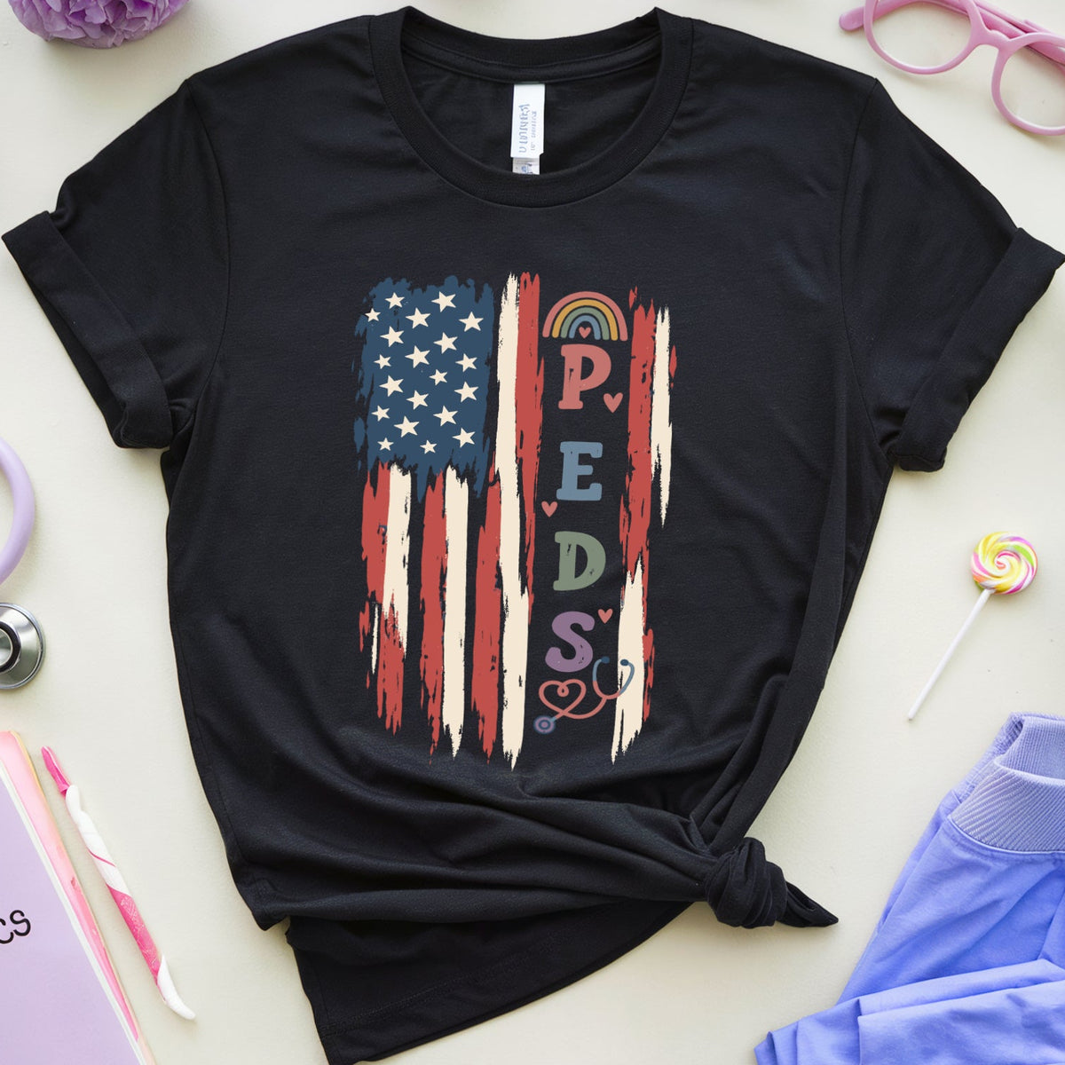 A black short-sleeve T-shirt with a distressed American flag graphic and the word 'PEDIATRICS' in colorful letters, styled in a flat lay with lavender flowers, a stethoscope, pink glasses, a notebook, and blue scrubs on a light beige background.
