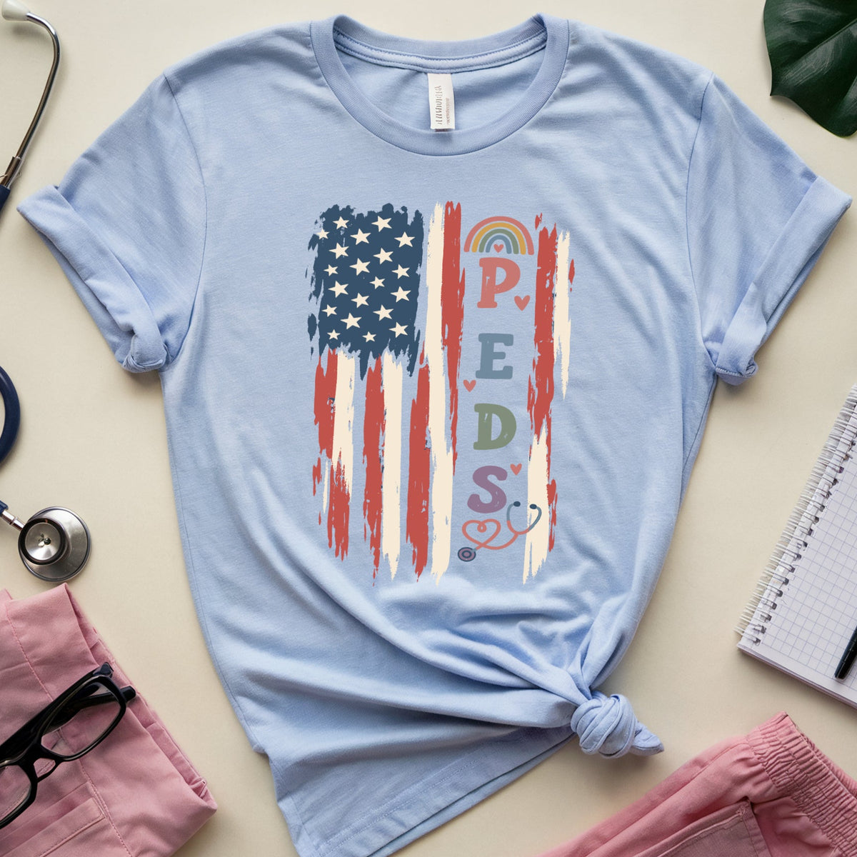 A light blue short-sleeve T-shirt laid flat on a cream surface, styled with a stethoscope, pink scrubs, glasses, a notebook, and a plant leaf. The shirt has a distressed American flag graphic with the letters 'P.