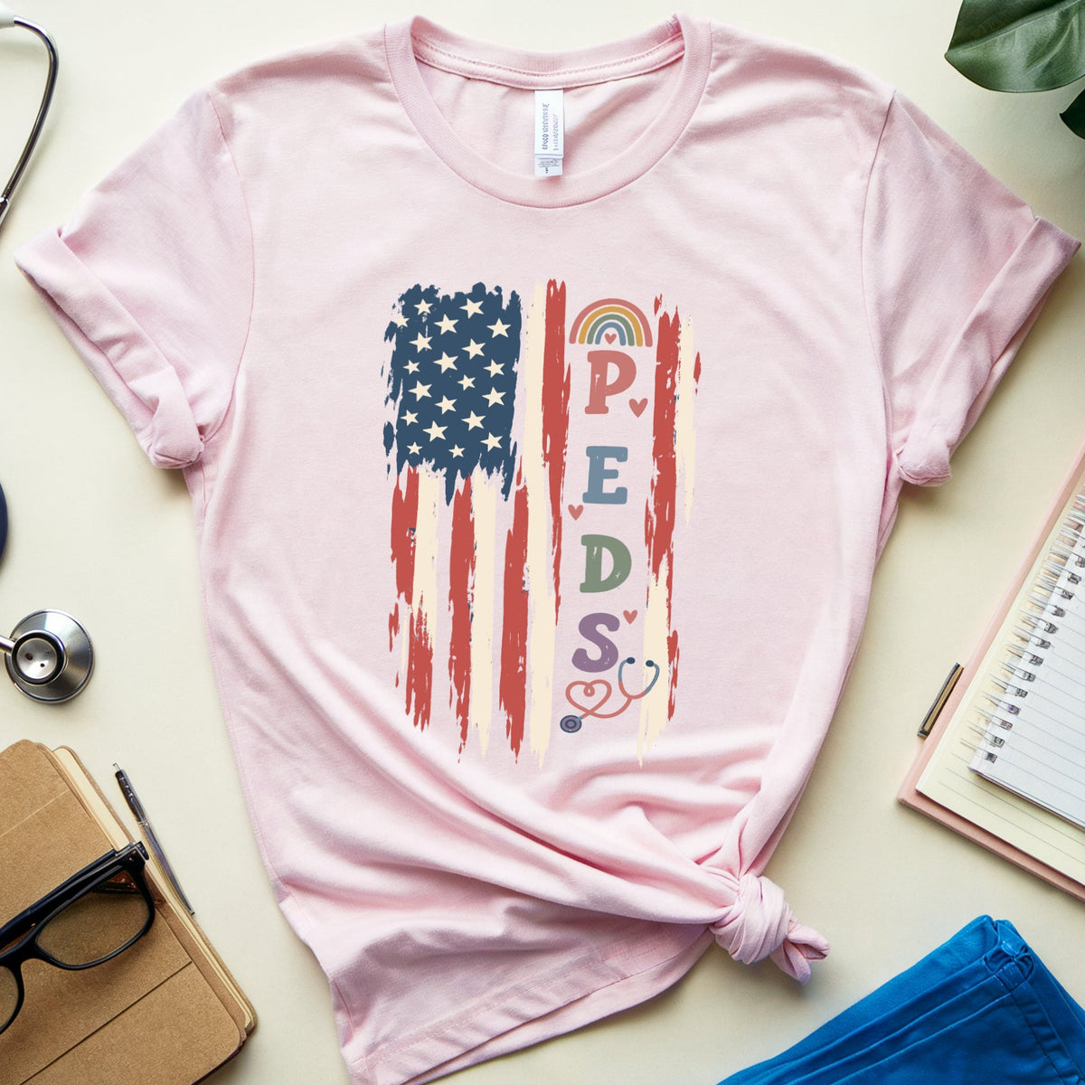 A light pink short-sleeve T-shirt laid flat on a cream surface, styled with a distressed American flag graphic and the word 'PROFESSIONAL' in rainbow colors. Surrounding props include a stethoscope, notebooks, glasses, a plant leaf, and blue pants.