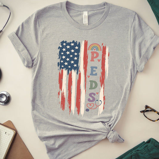A heather gray short-sleeve T-shirt laid flat on a light beige surface, styled with a knotted hem and surrounded by medical-themed props including a stethoscope, notebook, glasses, folded teal scrubs, and a small heart. The shirt features a distressed American flag graphic with the word 'PREDATORS' integrated into the stripes and stars.