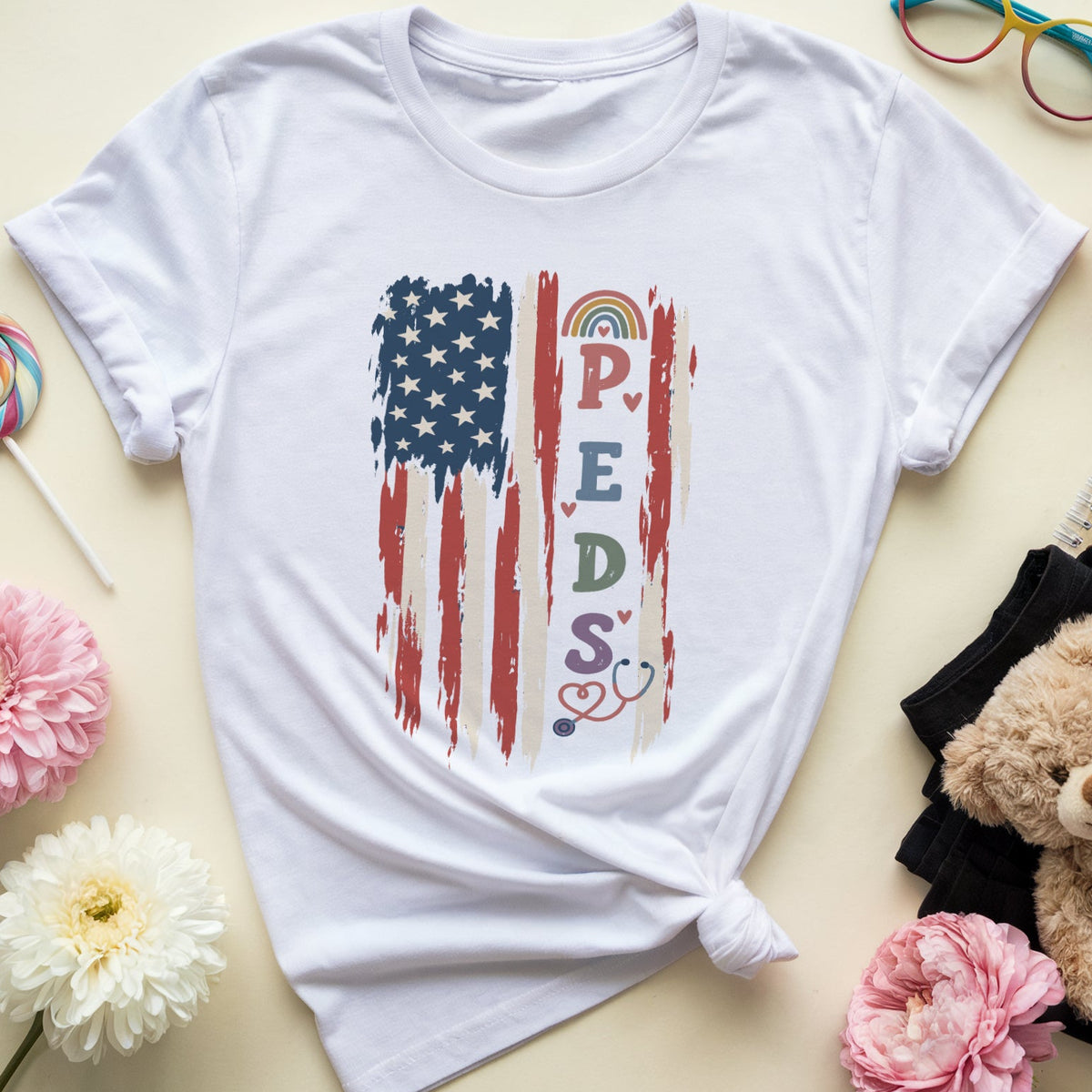 A white short-sleeve T-shirt laid flat on a light beige background, styled with flowers, glasses, a lollipop, a teddy bear, and a notebook. The shirt has a distressed American flag graphic with the word 'PEDS' and small heart and rainbow details.