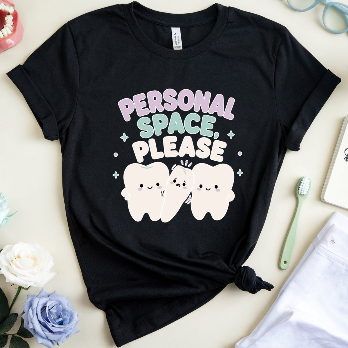 A black graphic t-shirt with a cute cartoon tooth design and the text 'PERSONAL SPACE, PLEASE' in pastel colors, styled in a flat lay with dental-themed props like a toothbrush, notebook, glasses, and a model teeth.