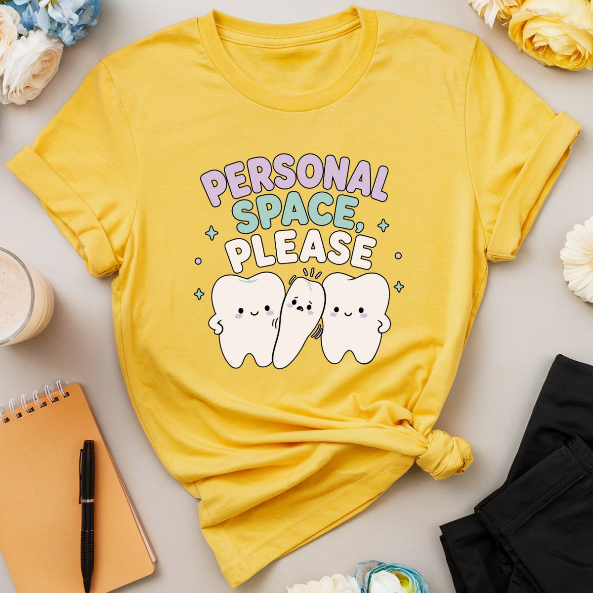 A yellow graphic T-shirt laid flat on a light beige surface, styled with flowers, glasses, a notebook, a pen, a drink, and black pants. The shirt has a colorful 'PERSONAL SPACE PLEASE' text design with three cartoon teeth characters.