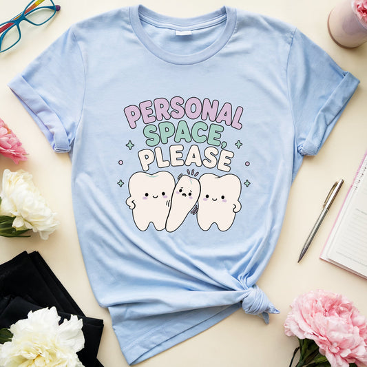 A light blue graphic t-shirt with a 'Personal Space Please' cartoon tooth design, styled in a flat lay with flowers, glasses, a notebook, and a pen on a cream background.