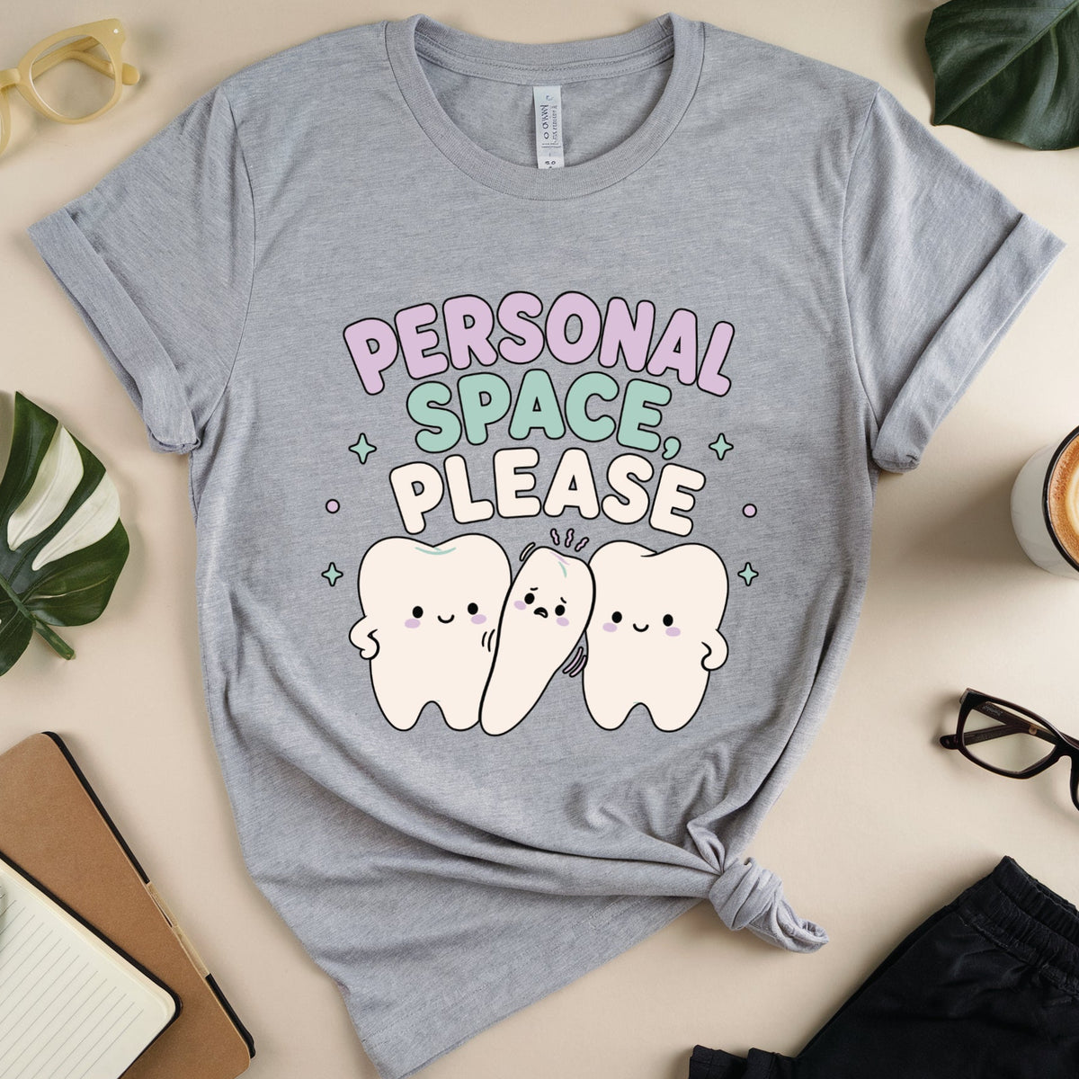 A heather gray graphic t-shirt with a 'Personal Space Please' cartoon tooth design, styled in a flat lay with glasses, a notebook, a coffee cup, and plant leaves on a beige background.