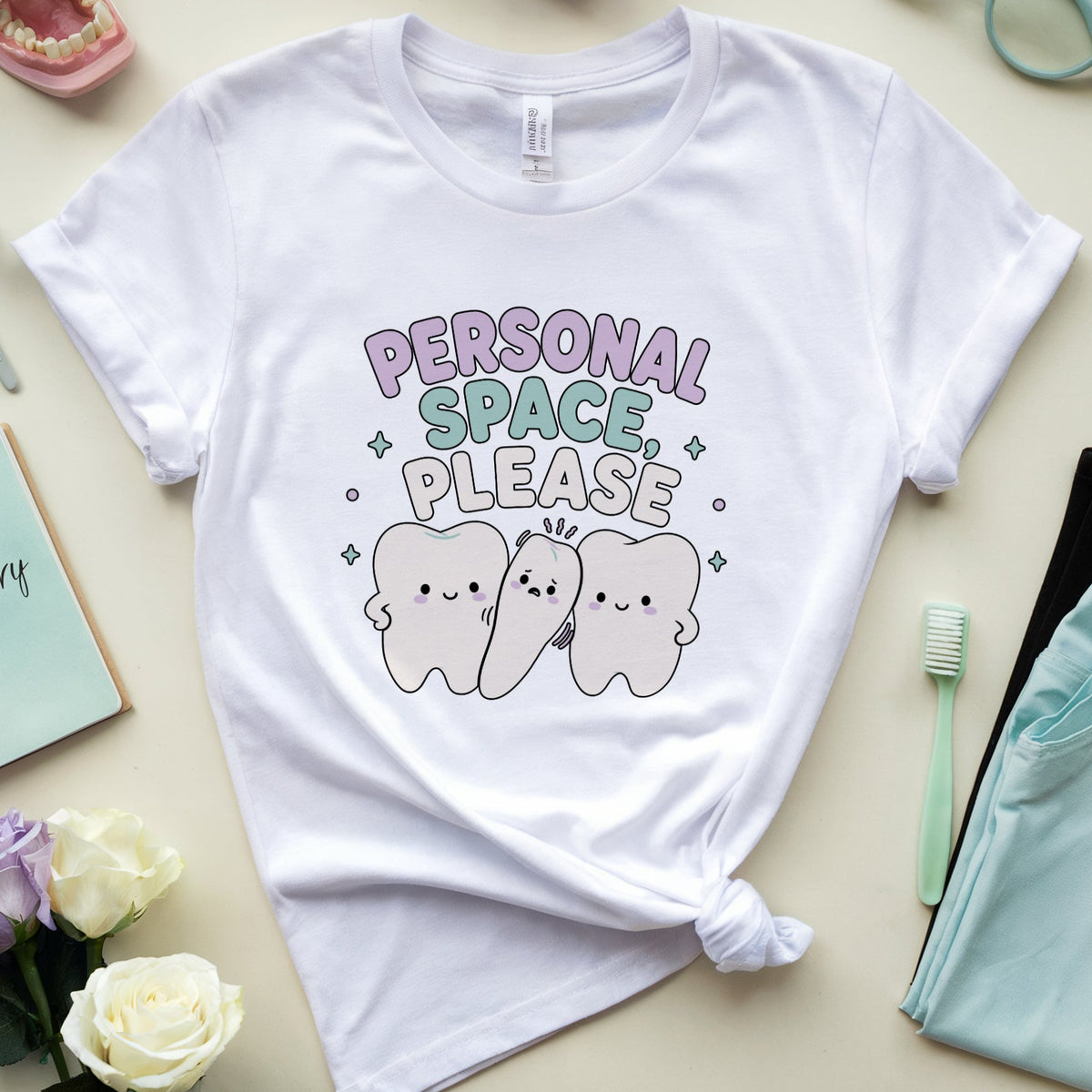 A white short-sleeve T-shirt laid flat on a light beige surface, styled with dental-themed props including a toothbrush, folded teal scrubs, a notebook, flowers, glasses, and a model mouth. The shirt has a printed graphic reading “PERSONAL SPACE PLEASE” above three cartoon teeth.
