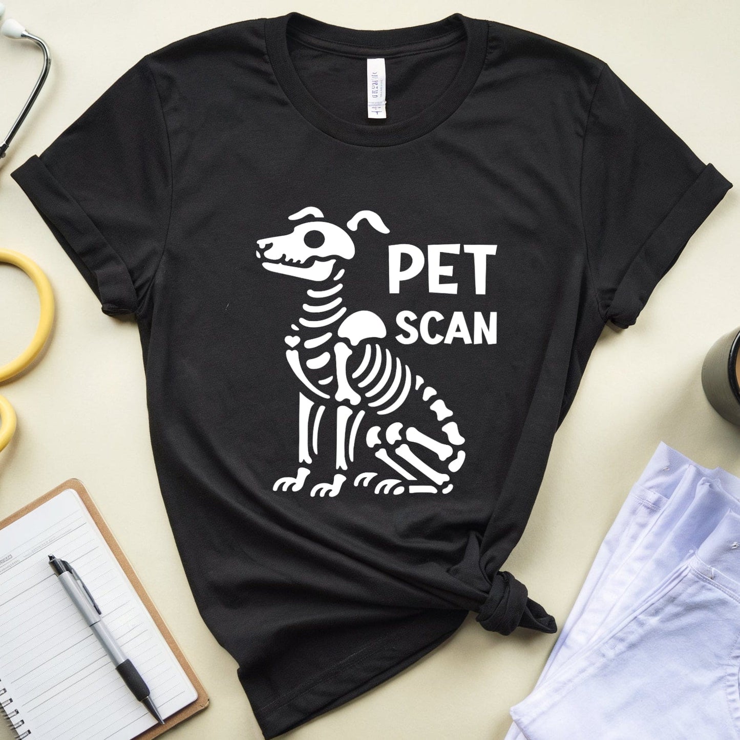 Black t-shirt with a dog skeleton graphic and 'PET SCAN' text on a light surface with medical-themed items.