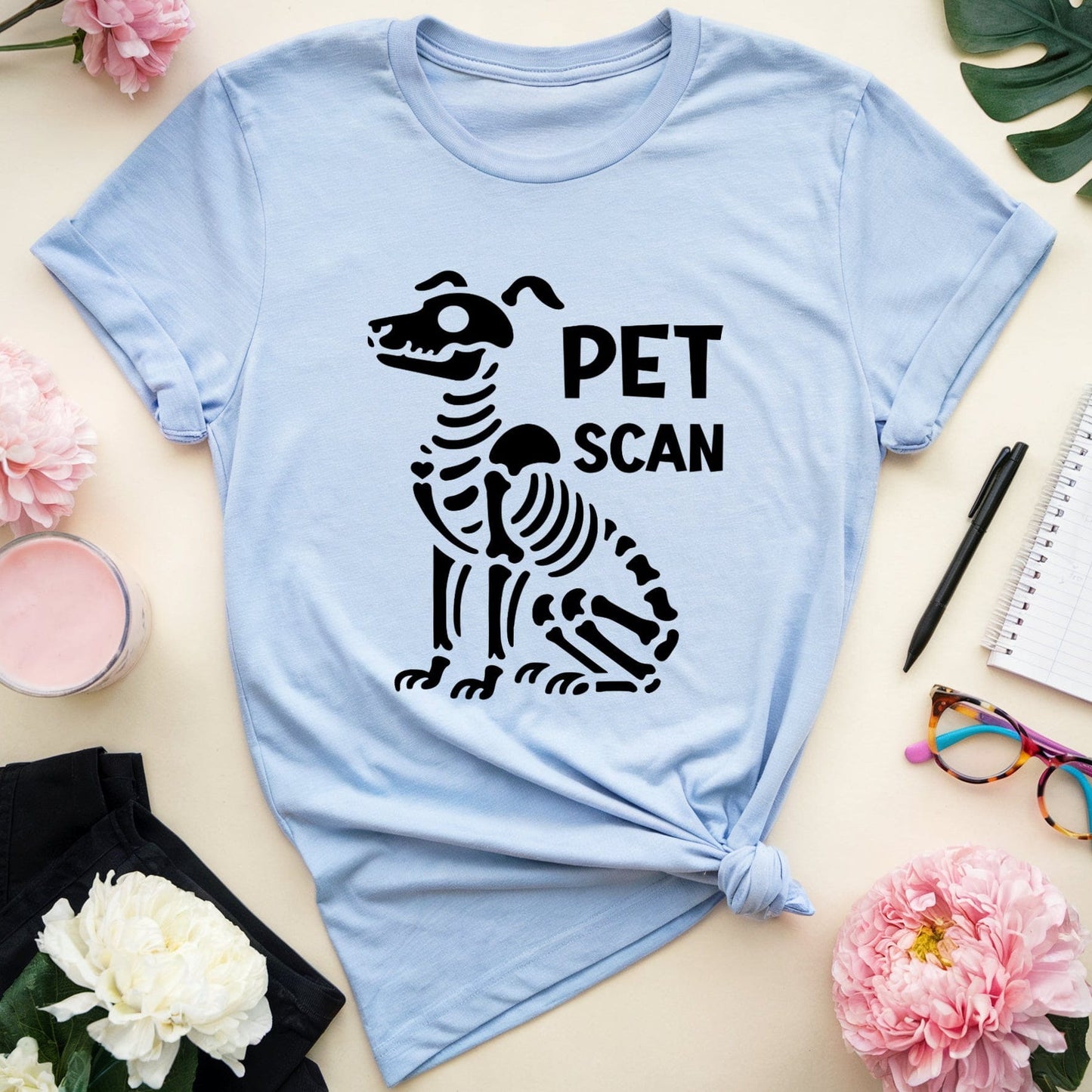 Light blue t-shirt with a black skeleton dog graphic and 'PET SCAN' text, placed on a surface with flowers and stationery.