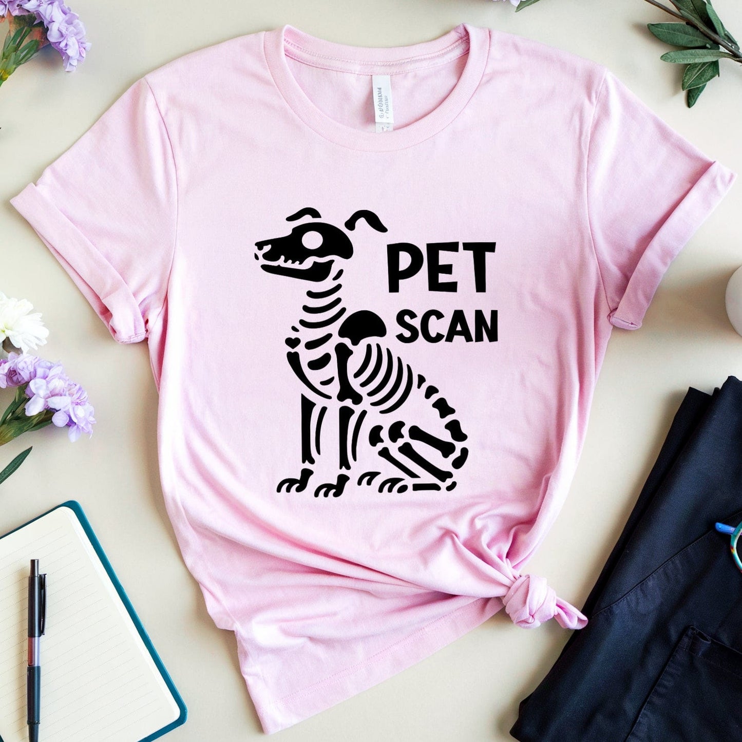 Pink t-shirt with a black skeleton design and 'PET SCAN' text, placed on a surface with flowers and a cup.