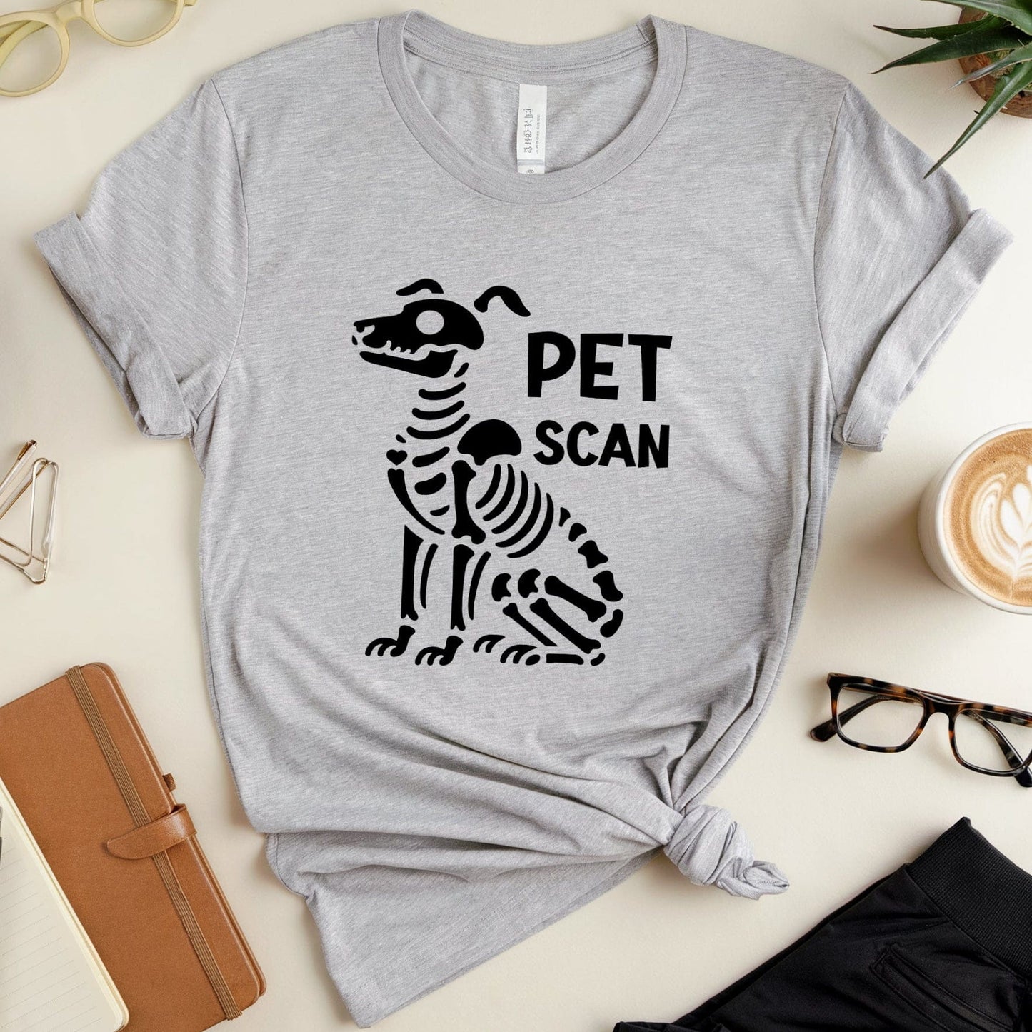 Gray t-shirt with a skeleton dog graphic and 'PET SCAN' text on a desk with office items.