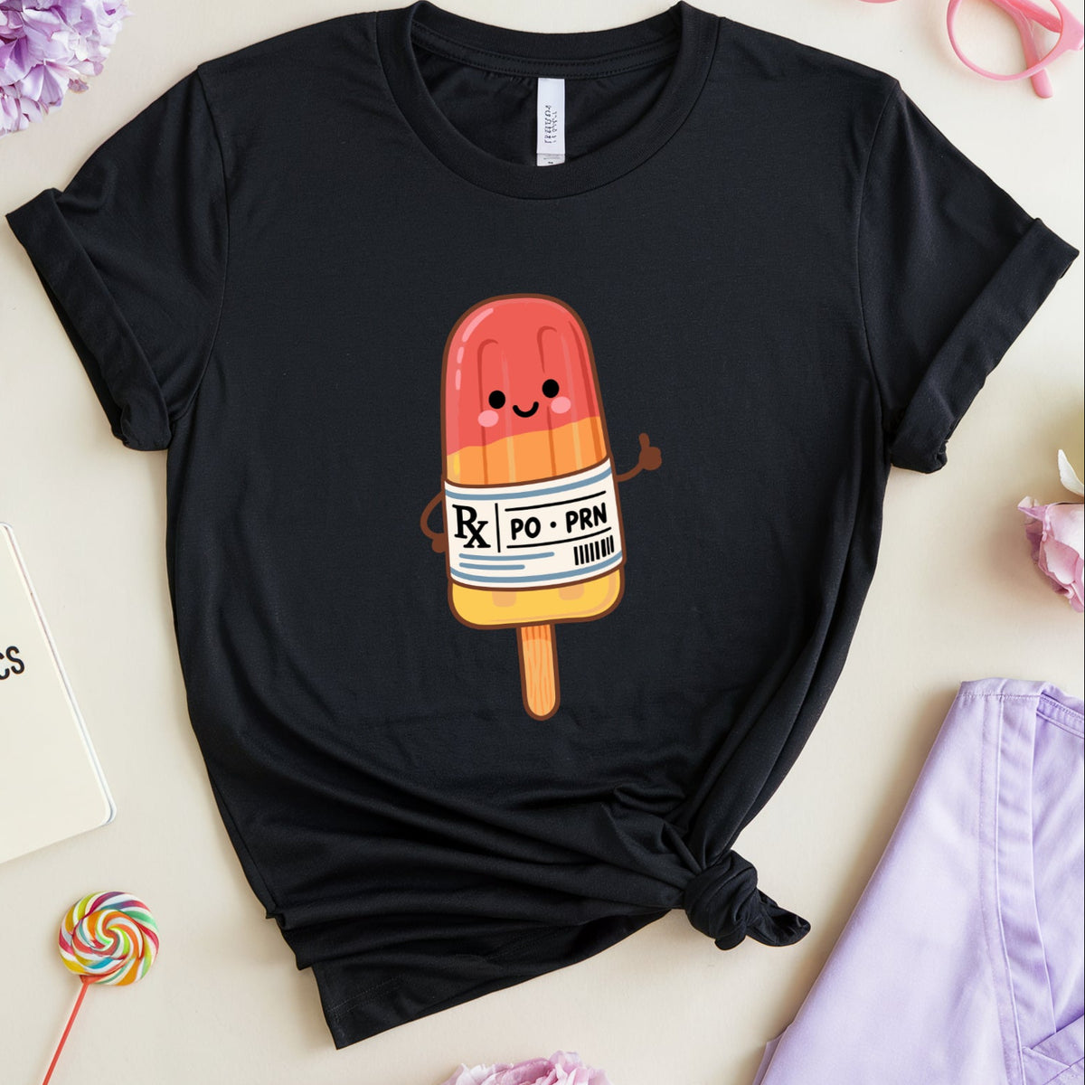 A black short-sleeve T-shirt laid flat on a light beige surface, styled with flowers, pink glasses, a notebook, a lollipop, and lavender scrubs. The shirt has a cartoon popsicle graphic on the chest with a red-orange top section, yellow-orange bottom section, and a white label reading 'Rx PO - POX'.