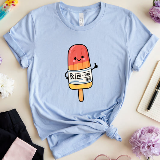 A light blue graphic t-shirt featuring a cute cartoon popsicle character with a 'POPO' label, styled in a flat lay with flowers, glasses, a notebook, and pants on a light beige background.