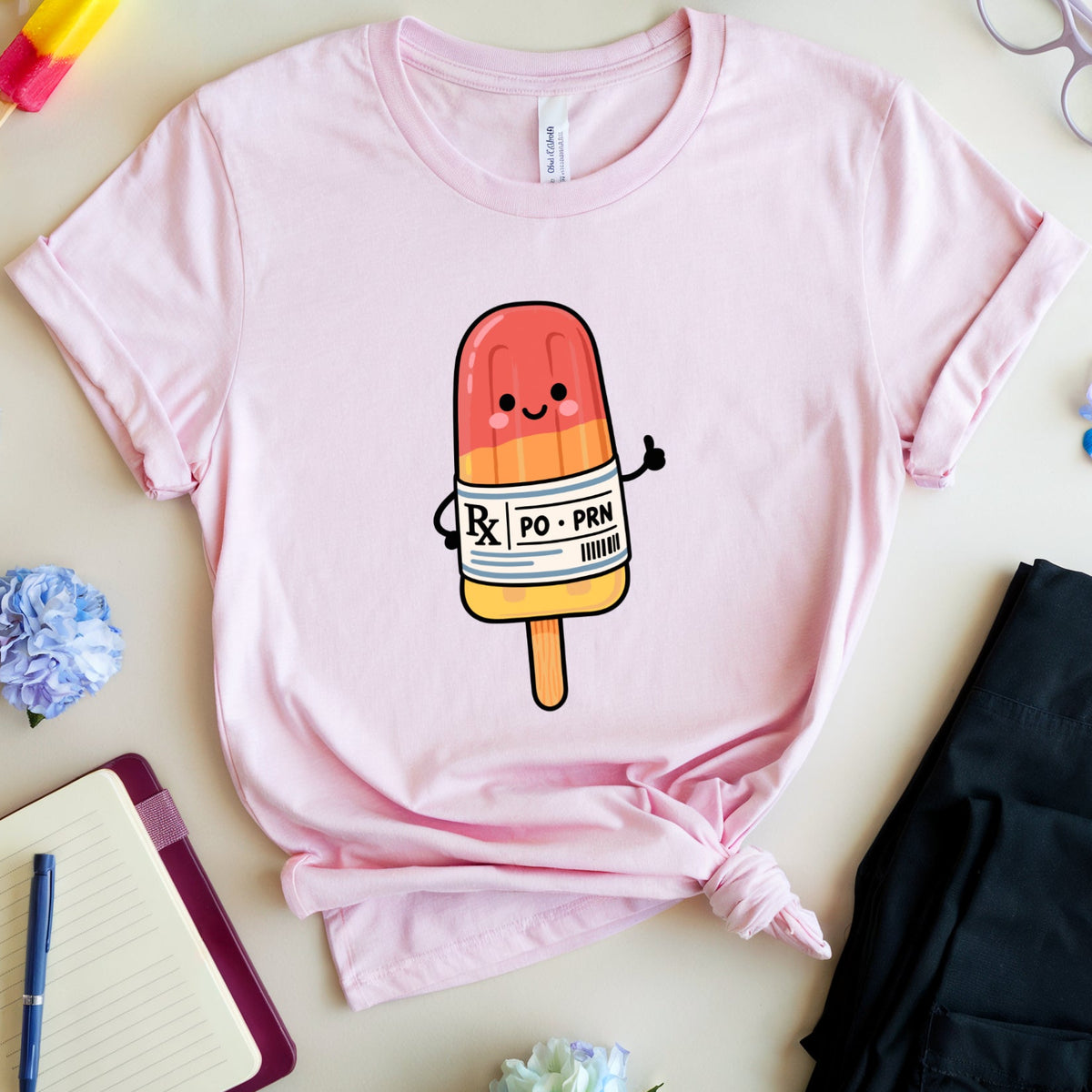 A light pink short-sleeve T-shirt laid flat on a light beige surface, styled with a tie-front knot and a cute popsicle graphic on the chest. The shirt is surrounded by flowers, glasses, a notebook, a pen, and folded black pants.