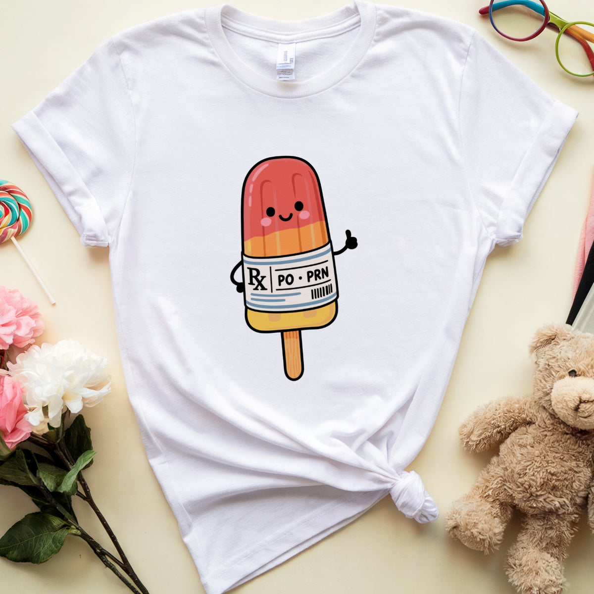 A white short-sleeve T-shirt laid flat on a light beige background, styled with flowers, glasses, a teddy bear, a book, and a lollipop. The shirt has a cartoon popsicle graphic on the chest with a pink-to-orange body, a white label reading 'POPSicle', and a wooden stick.