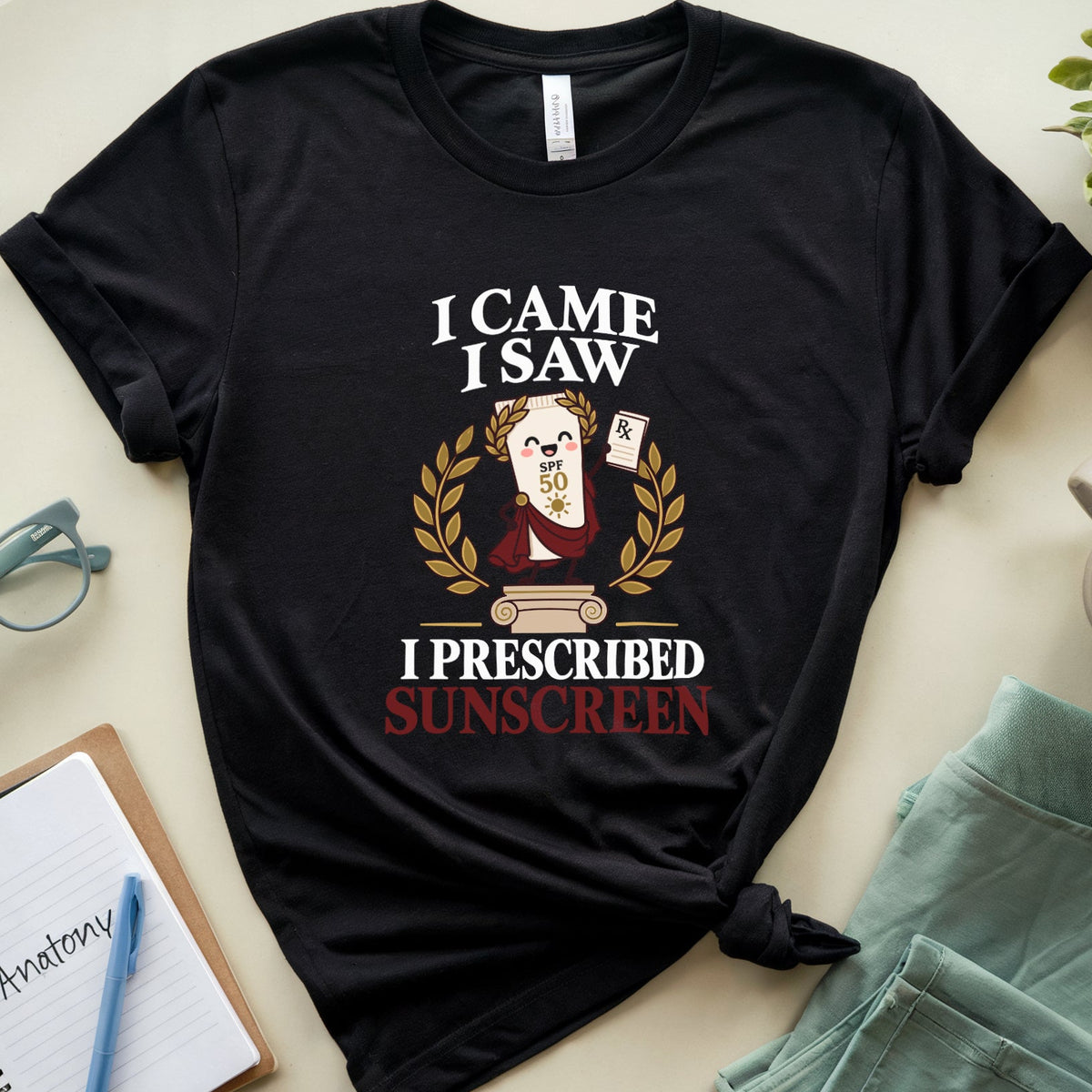 A black graphic t-shirt laid flat on a light beige surface, styled with a notebook, glasses, a mug, mint green shorts, and a small plant. The shirt features a cartoon sloth wearing a sun hat and sunglasses, surrounded by a laurel wreath, with the text 'I CAME I SAW I PRESCRIBED SUNSCREEN'.