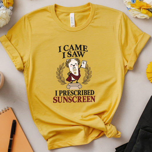 A yellow graphic t-shirt with a cartoon monkey wearing a sunscreen bottle and the text 'I CAME I SAW I PRESCRIBED SUNSCREEN'. The shirt is styled in a flat-lay arrangement with flowers, glasses, a notebook, and a pair of black pants on a light beige background.