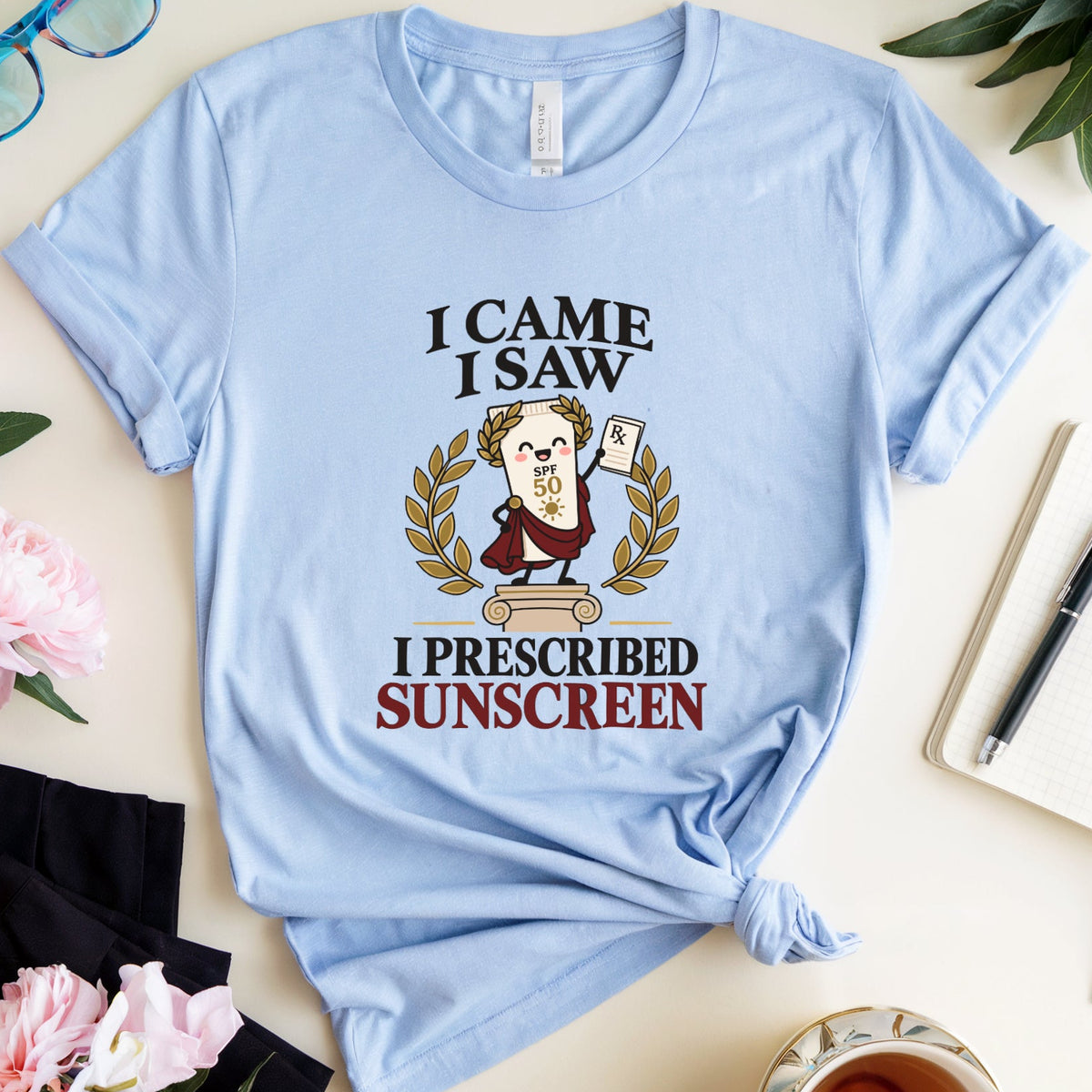 A light blue graphic t-shirt is displayed in a styled flat-lay arrangement on a light beige surface. The shirt features a cartoon illustration of a man with glasses and a sun hat, surrounded by a laurel wreath, with the text 'I CAME I SAW I PRESCRIBED SUNSCREEN'.